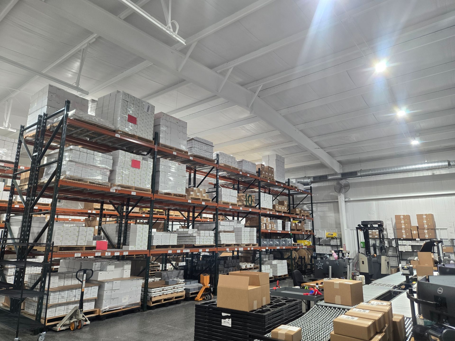 Warehouse interior with packed shelves and a forklift. Boxes and pallets line the floor. Bright overhead lights.