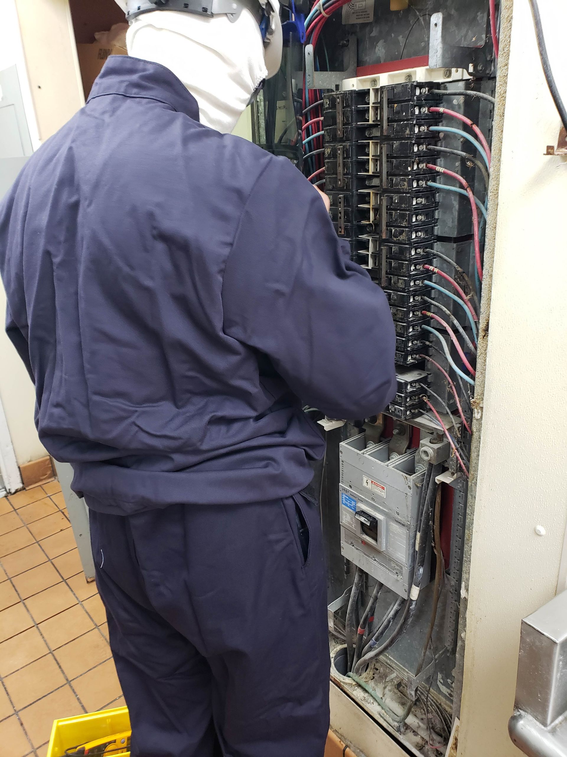 Electrician in blue uniform working inside an open electrical panel, examining wires.