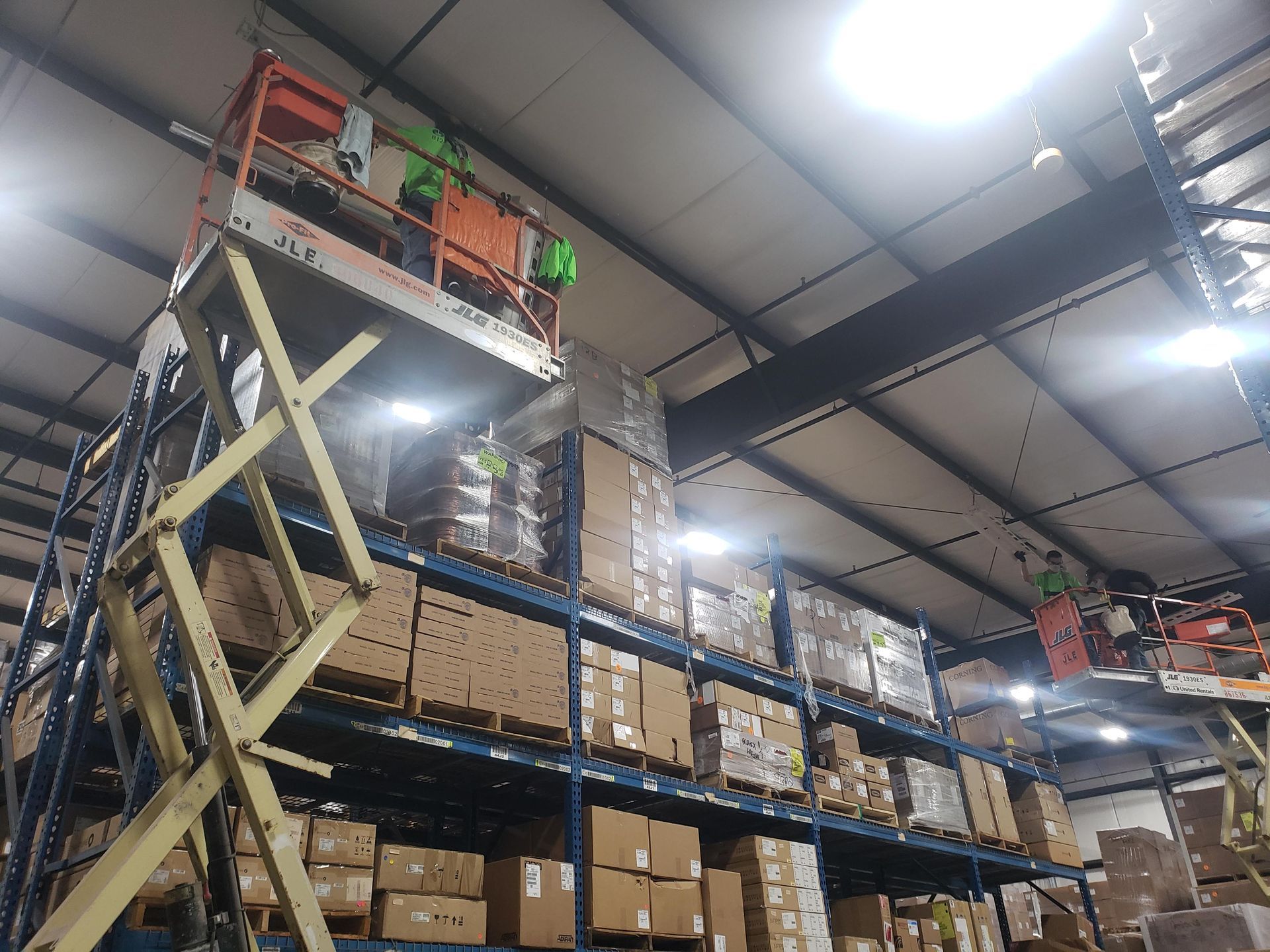 Warehouse workers on scissor lifts, accessing high shelves filled with boxes.
