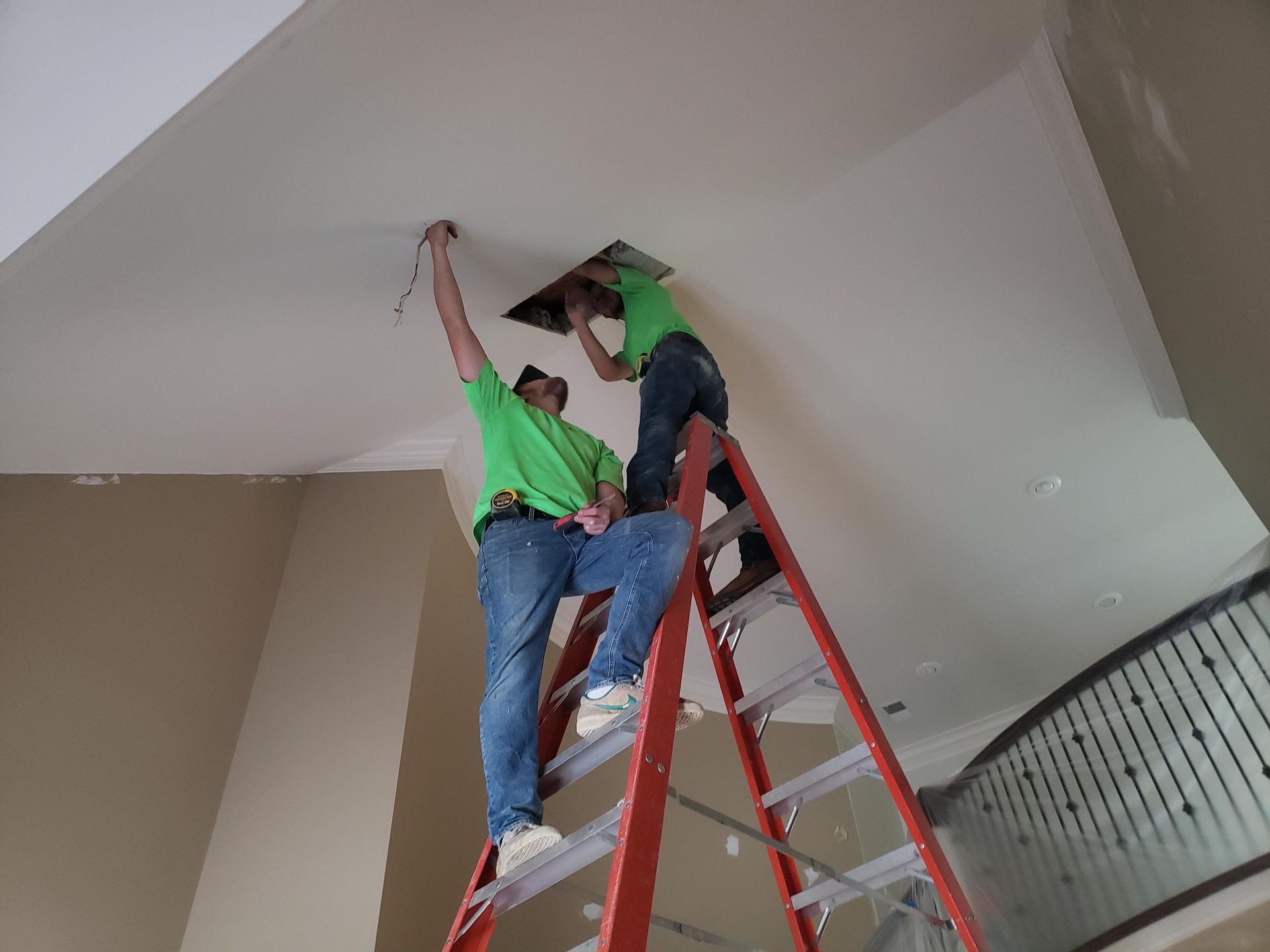 Two people on a ladder, working on ceiling. One holds wires, the other looks inside an opening.