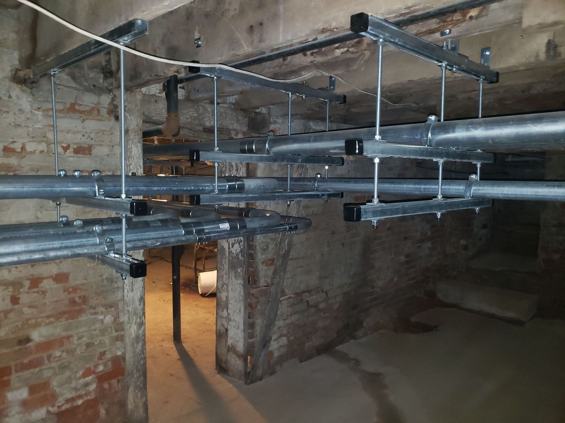 Pipes suspended from the ceiling in a brick basement. Gray metal framing supports the pipes.