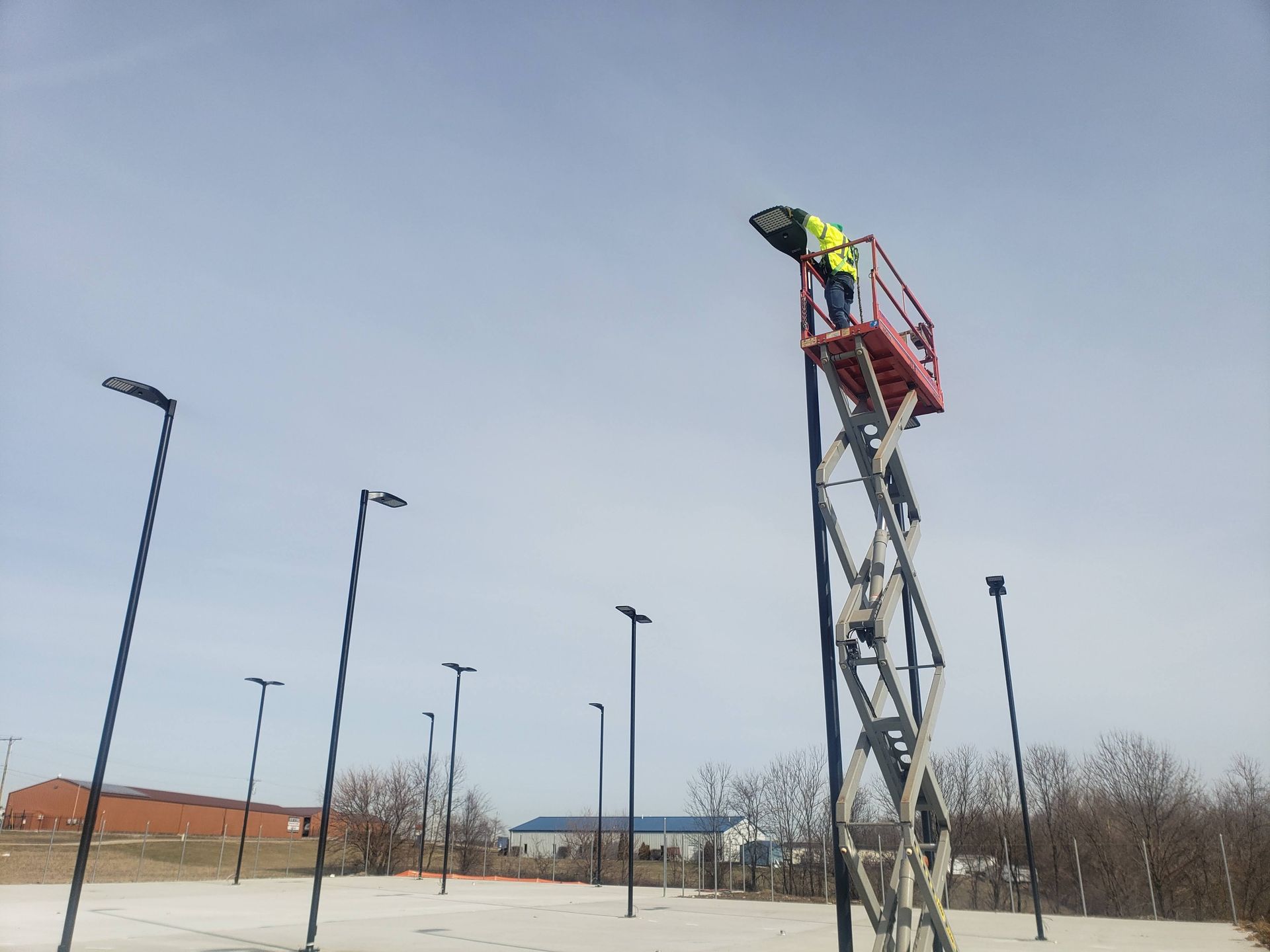 Person in safety vest on a lift, installing a light fixture on a pole. Other light poles are in the background. Blue sky.