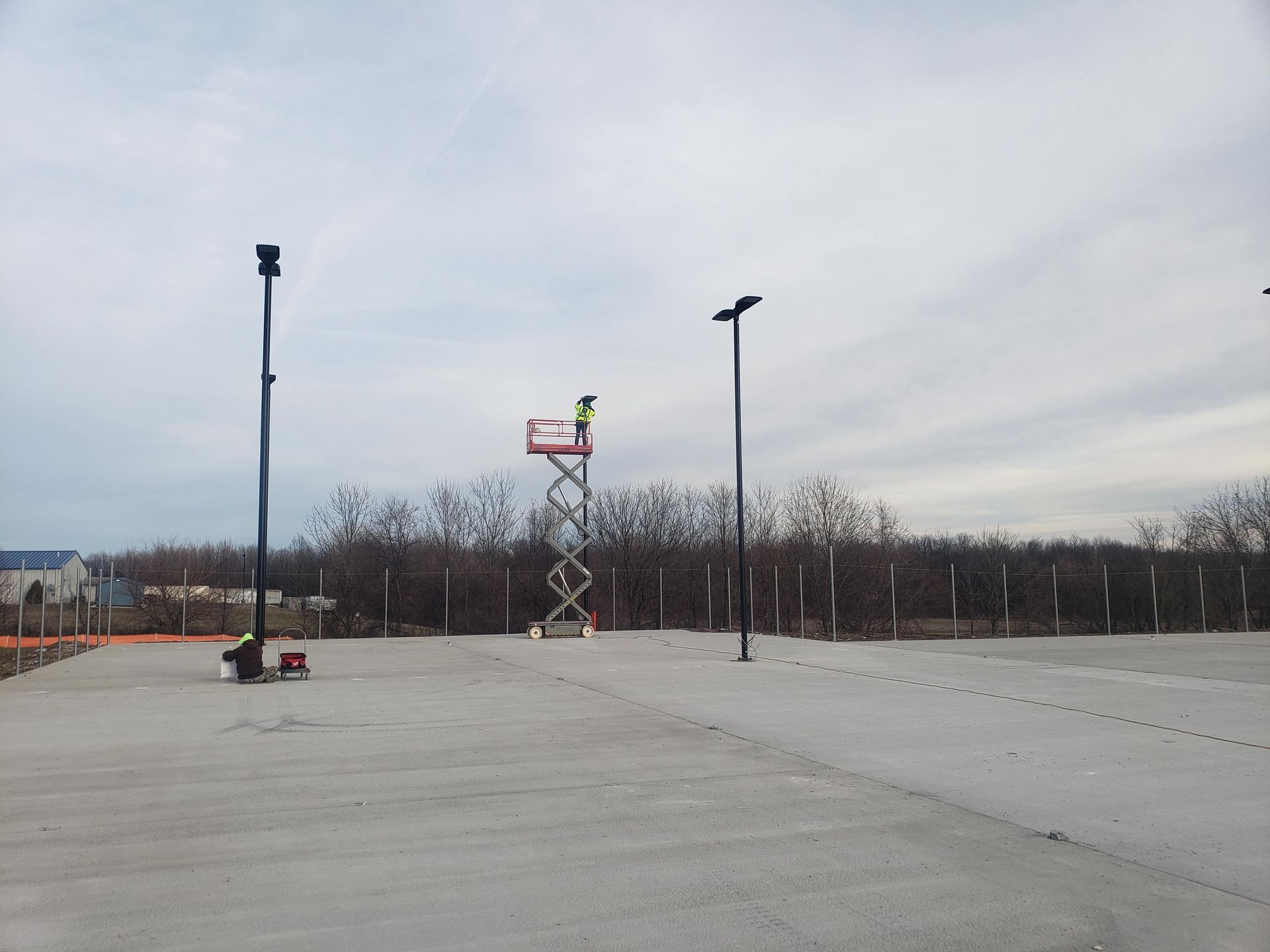 Person on a lift working on a light fixture outdoors on a concrete surface. Two other light poles stand nearby.