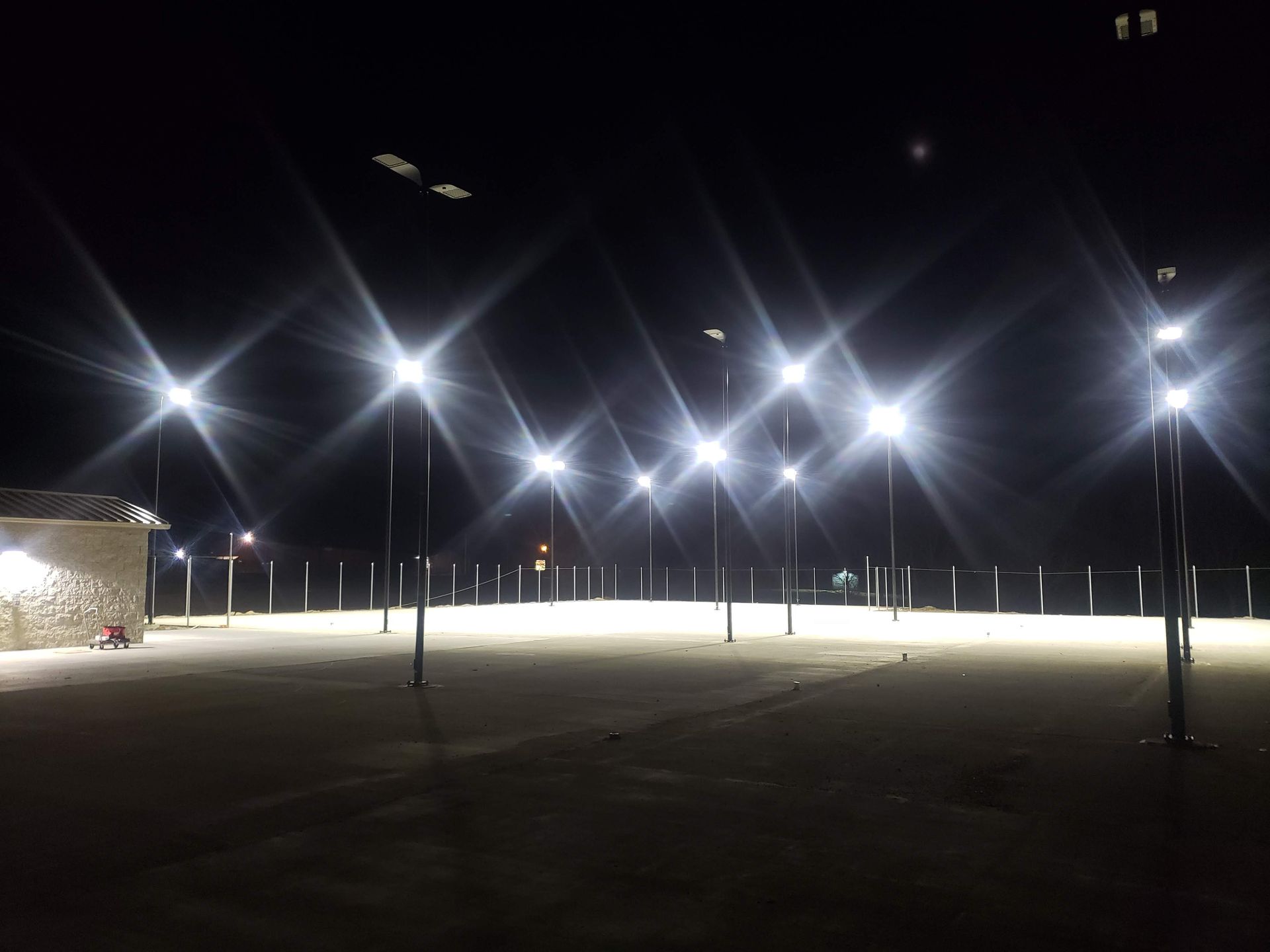 Nighttime view of a sports field illuminated by numerous bright light posts; building on the left, fence behind the field.
