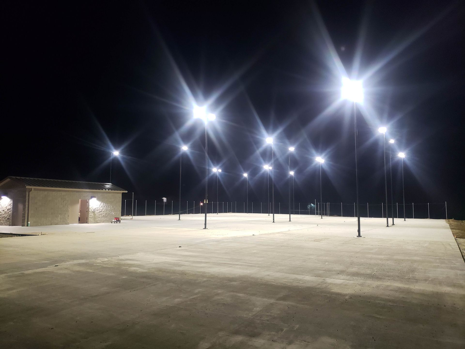 Nighttime view of a brightly lit parking lot with numerous tall light poles; a small building is on the left.