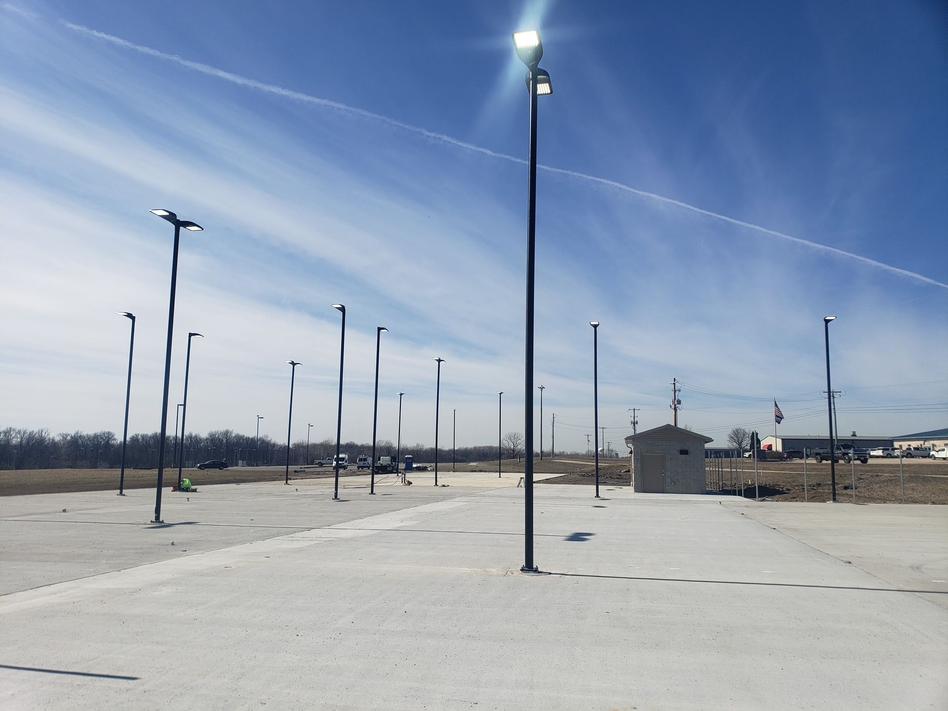 Parking lot with tall, dark light poles on a sunny day. A small concrete building sits in the background.