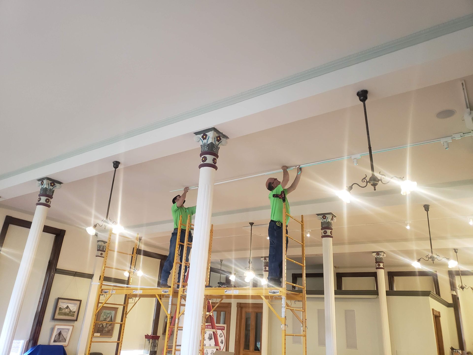 Two people on scaffolding installing trim on a ceiling, near decorative columns, inside a building.