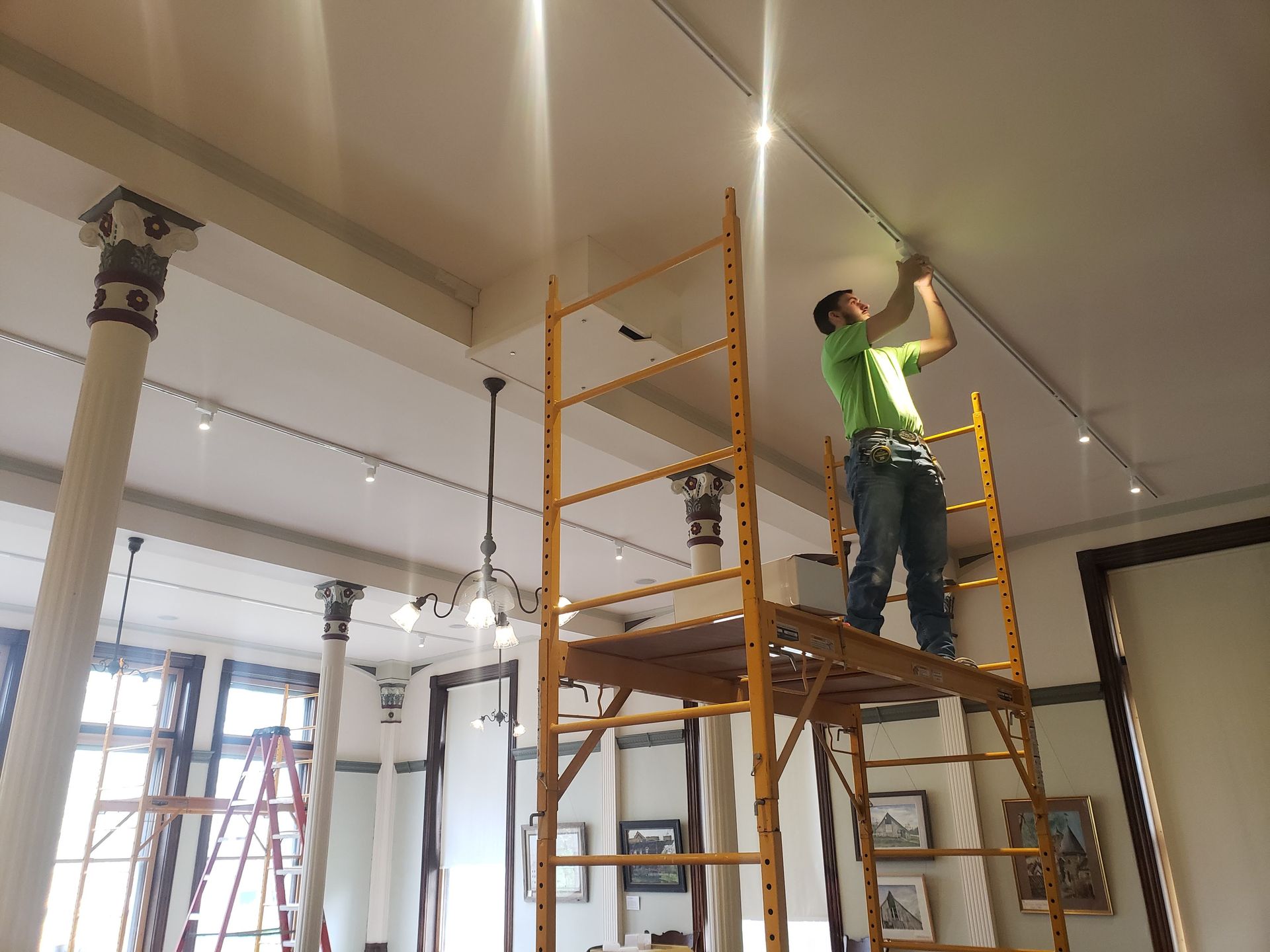 A person on scaffolding installing track lighting in a gallery with decorative columns.