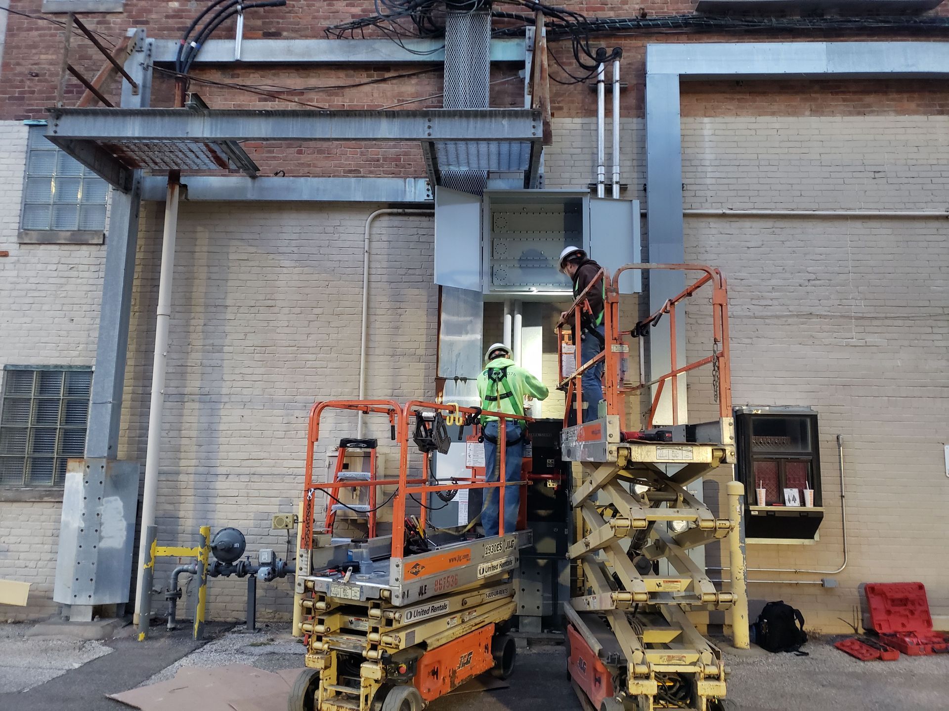 Construction workers on lifts installing electrical equipment on a brick building exterior.