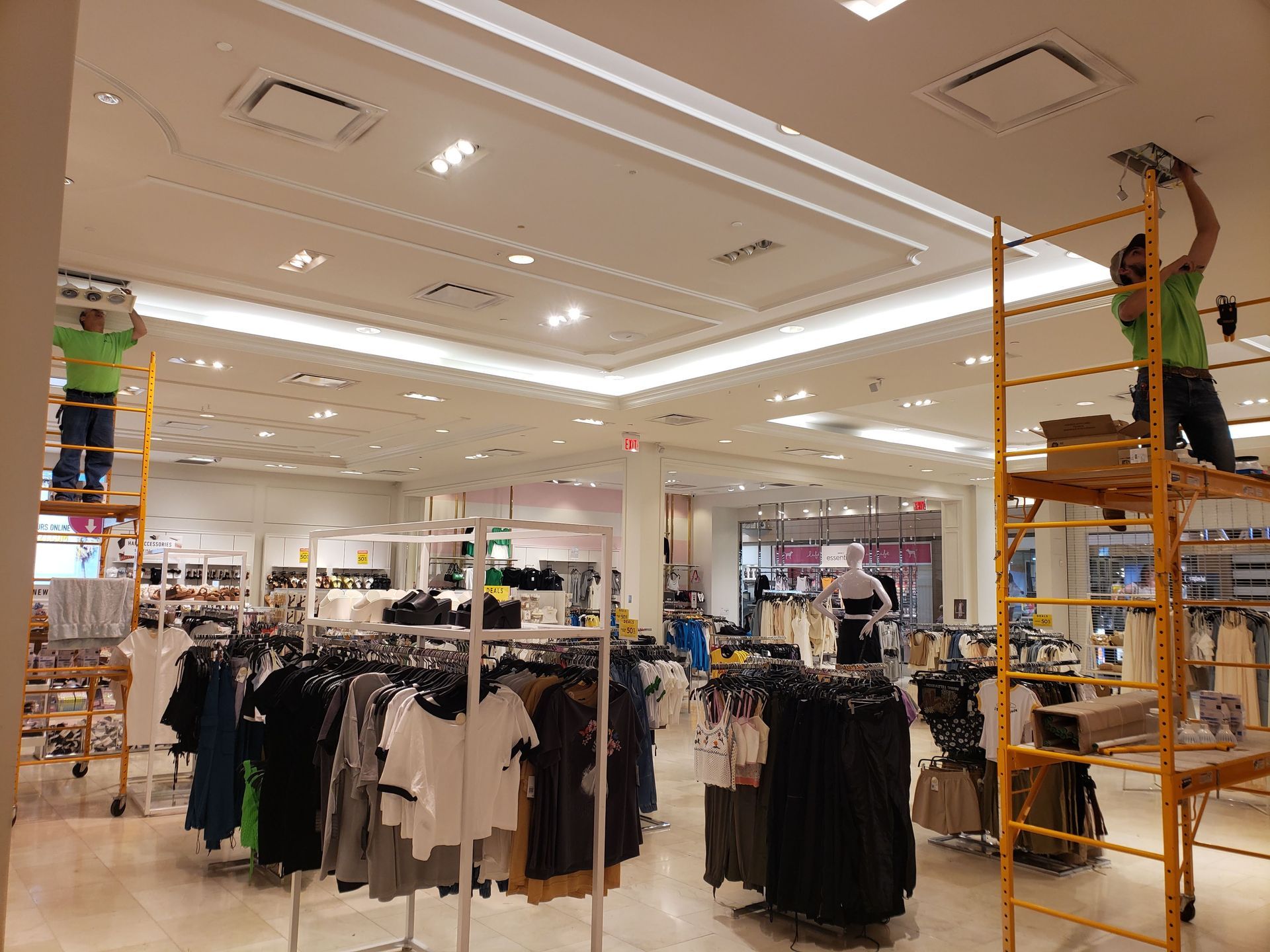 Two workers on scaffolding installing ceiling fixtures in a clothing store. Clothes racks fill the foreground.