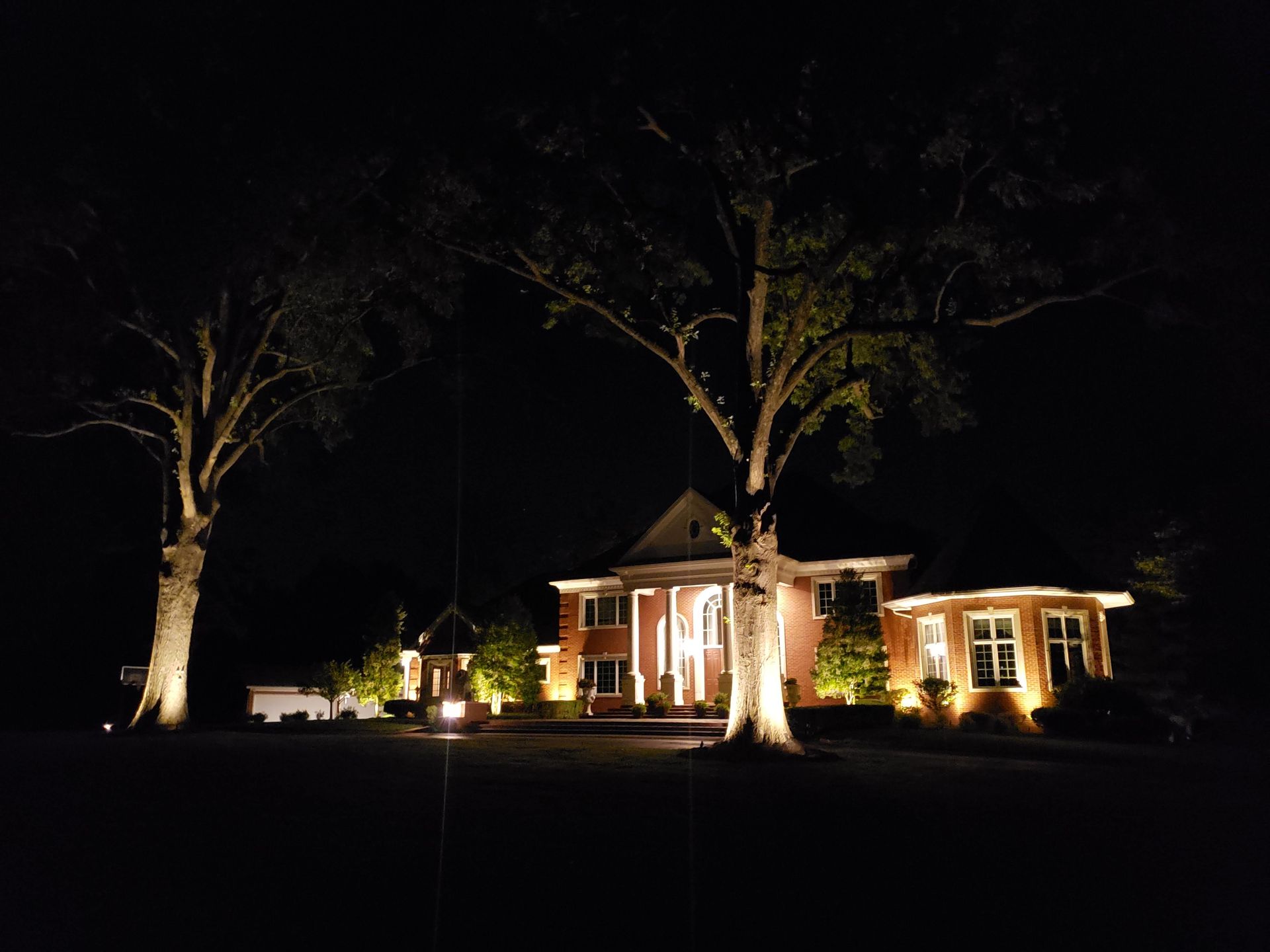 A large brick house at night, lit with outdoor lights. Two trees flank the house.
