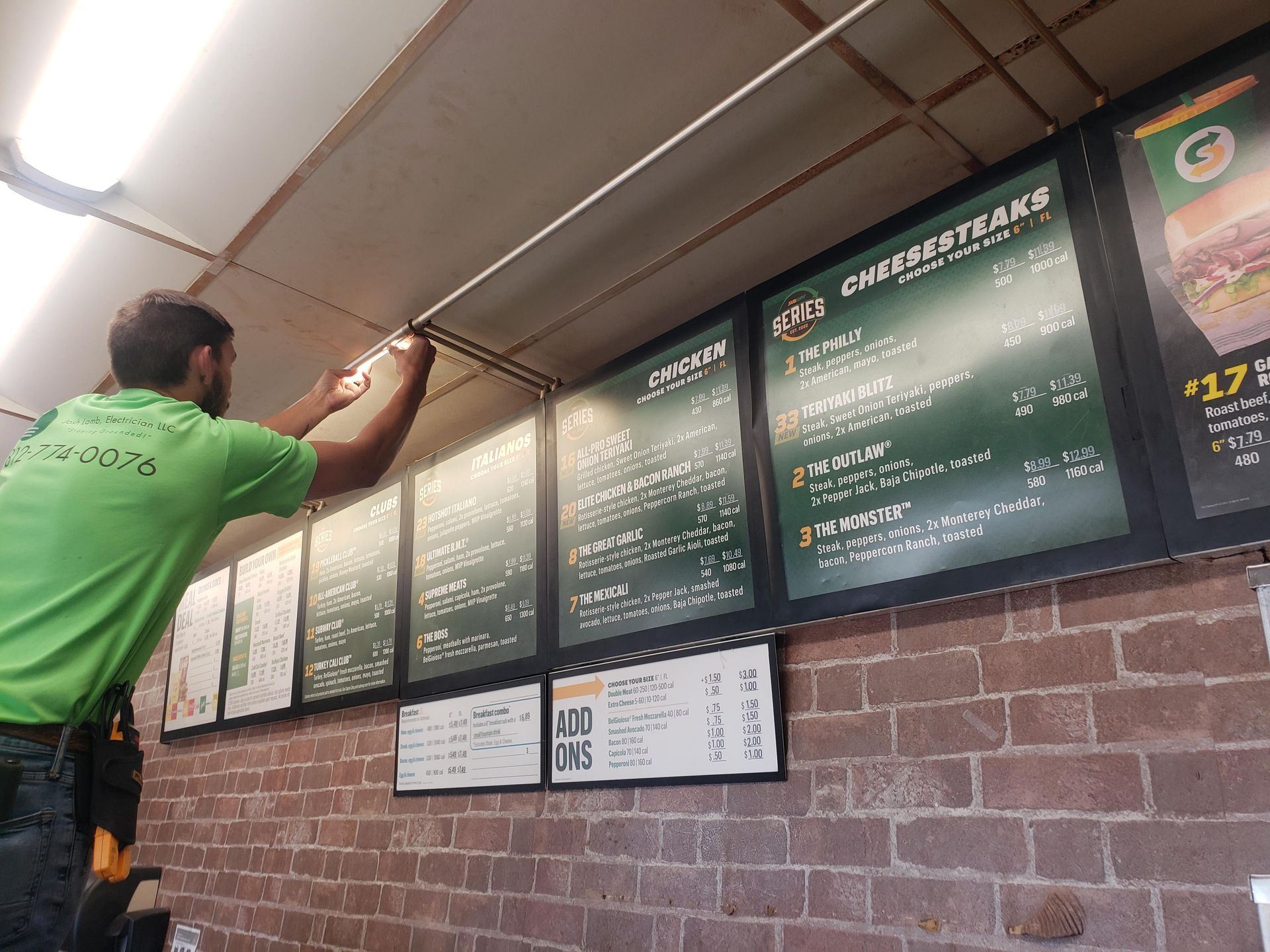 Man in green shirt installing lights above Subway menu boards on a brick wall.
