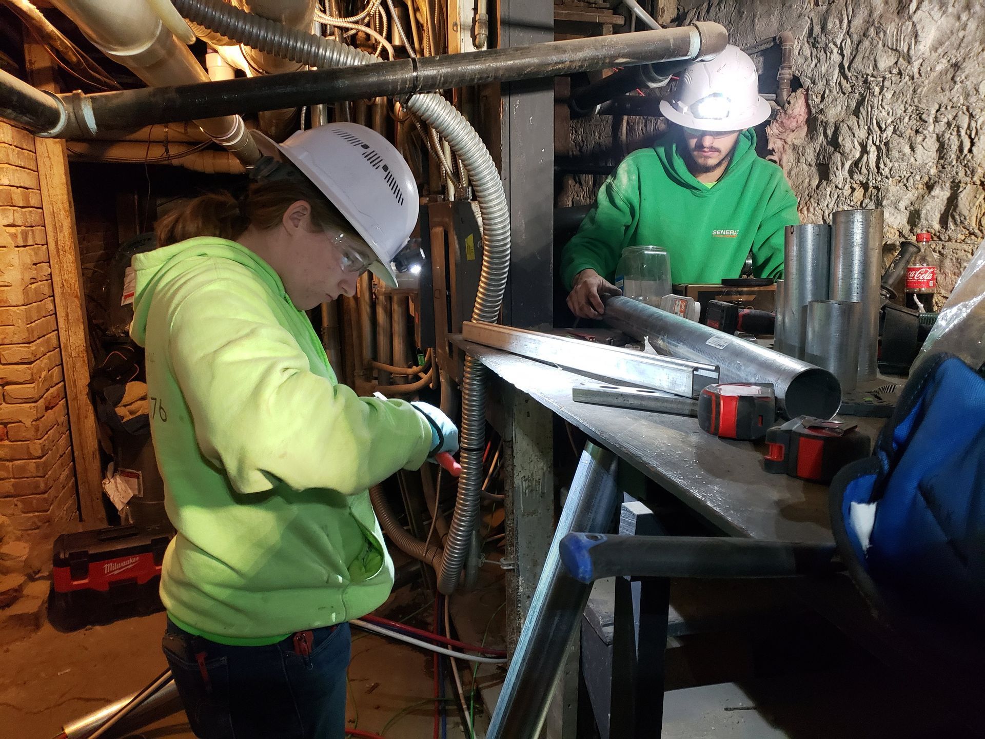 Two people in hard hats work on pipes in a dimly lit industrial setting. One cuts wire, the other works with metal.