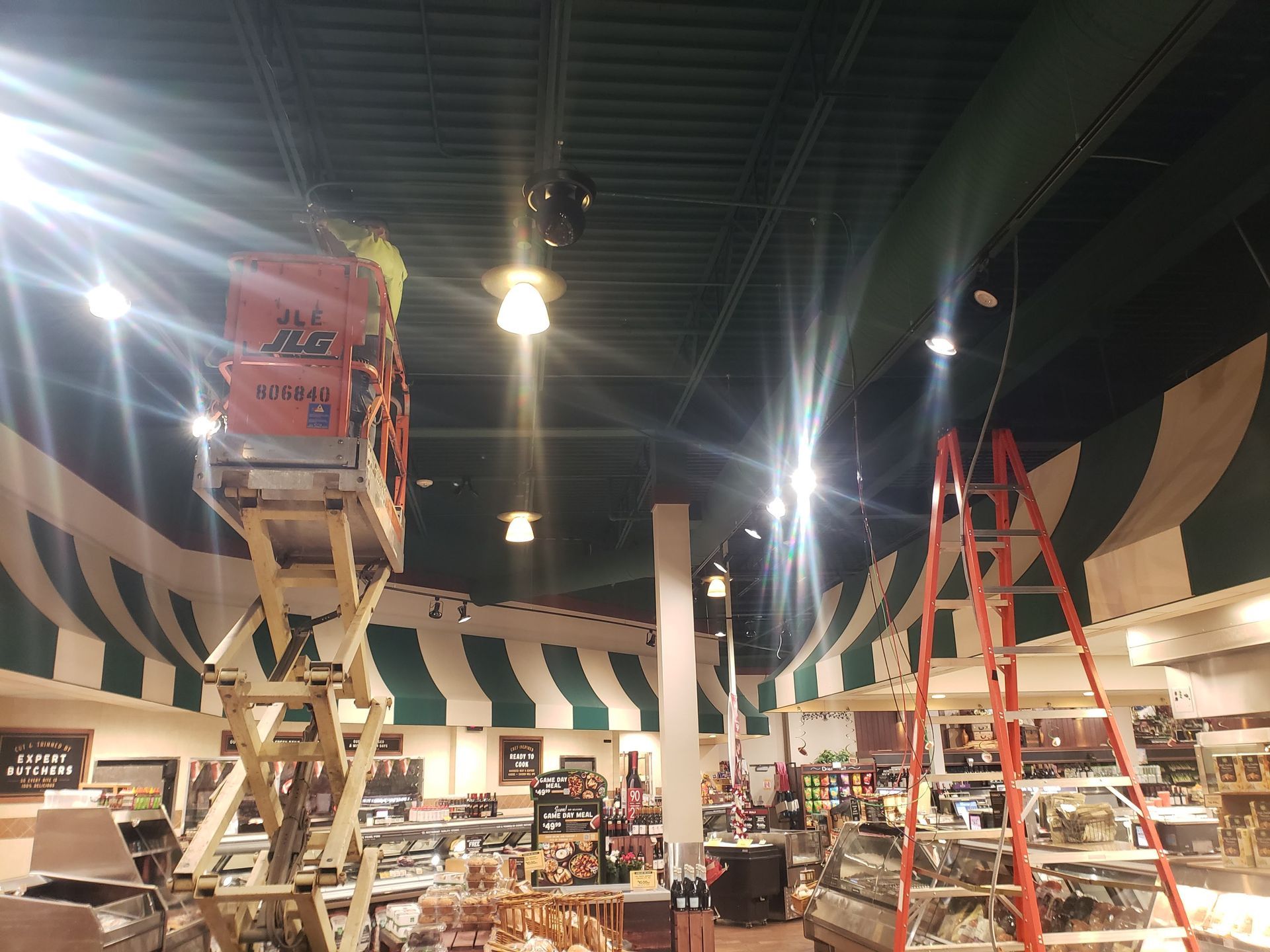 Person in a lift working on ceiling lights in a store. Ladders, green and white awning, and merchandise are visible.