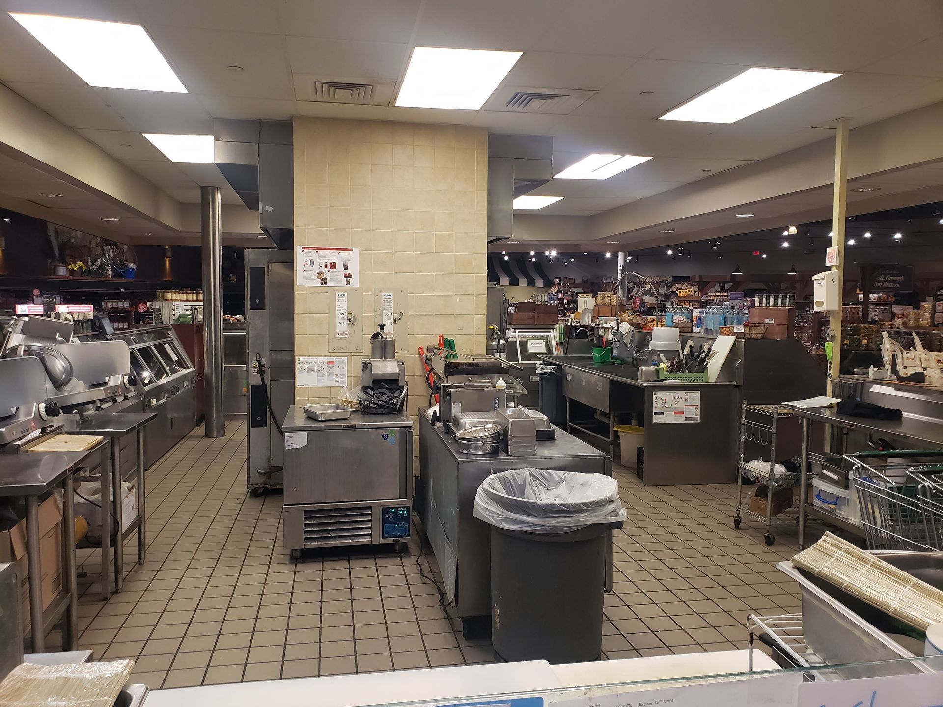 A commercial kitchen with stainless steel appliances and tile flooring. Fluorescent lights illuminate the space.