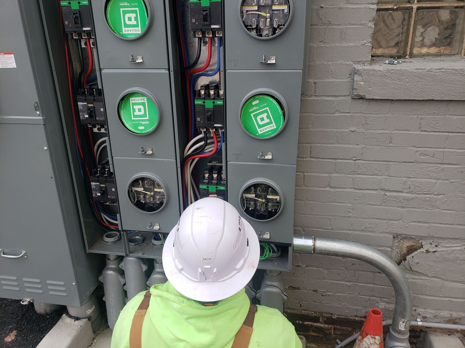 Person in hard hat examines electrical meters on a brick building exterior.