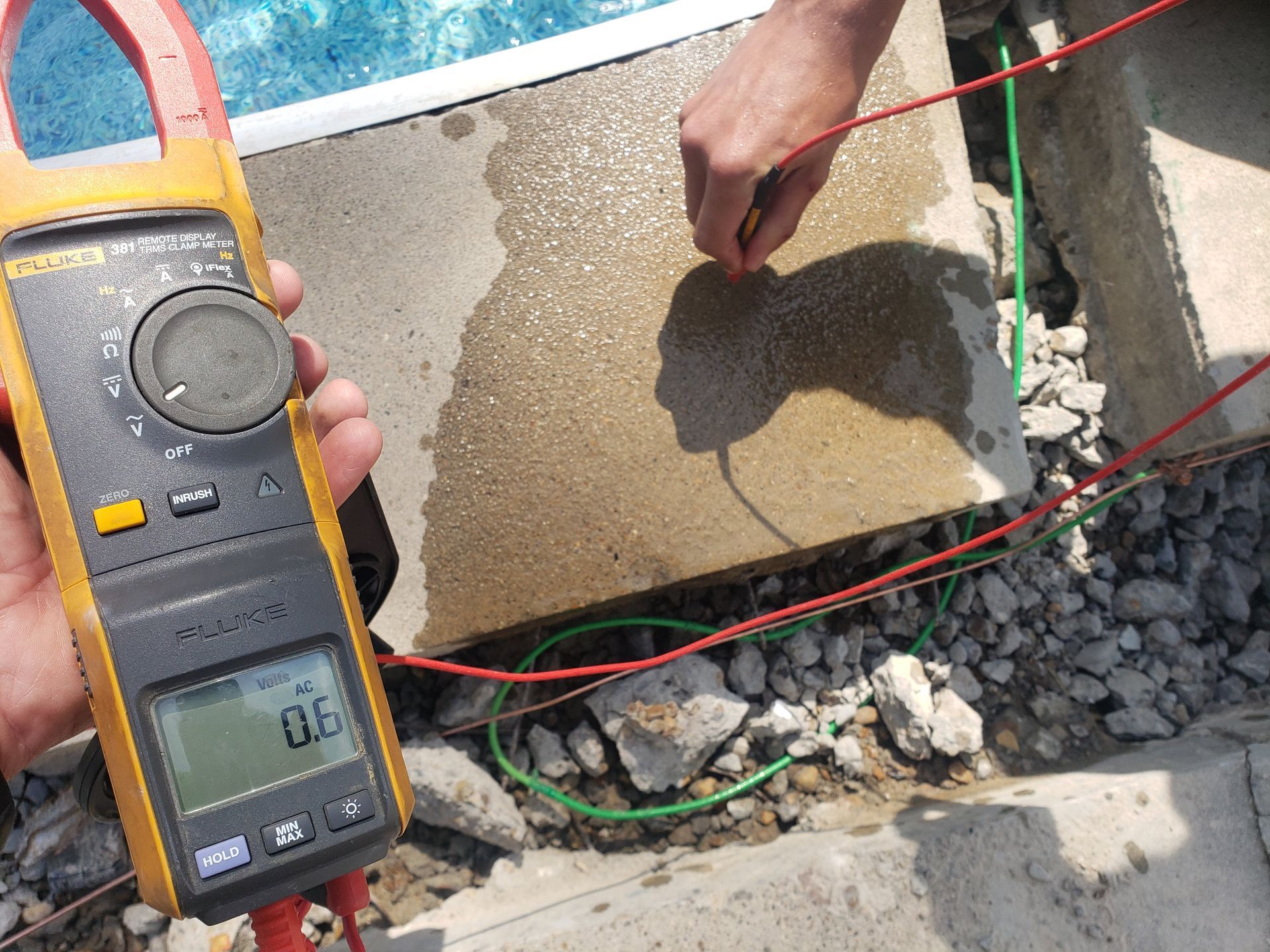 A person tests electrical current on wet concrete with a multimeter. Red and green wires are attached.