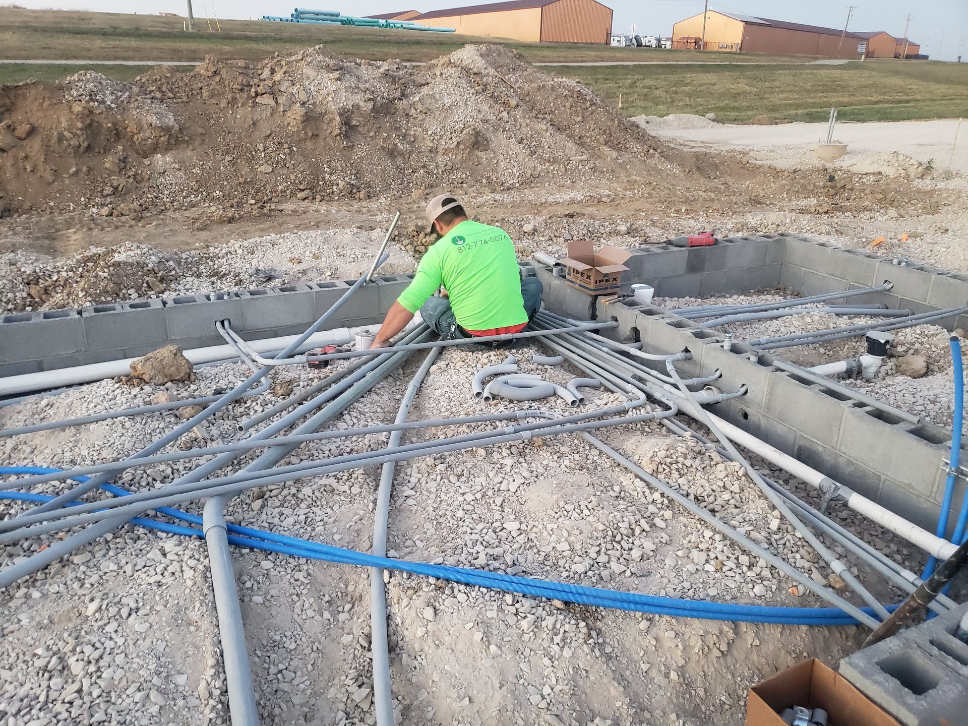 Construction worker installing electrical conduit within a foundation. Gravel, pipes, and concrete blocks are present.
