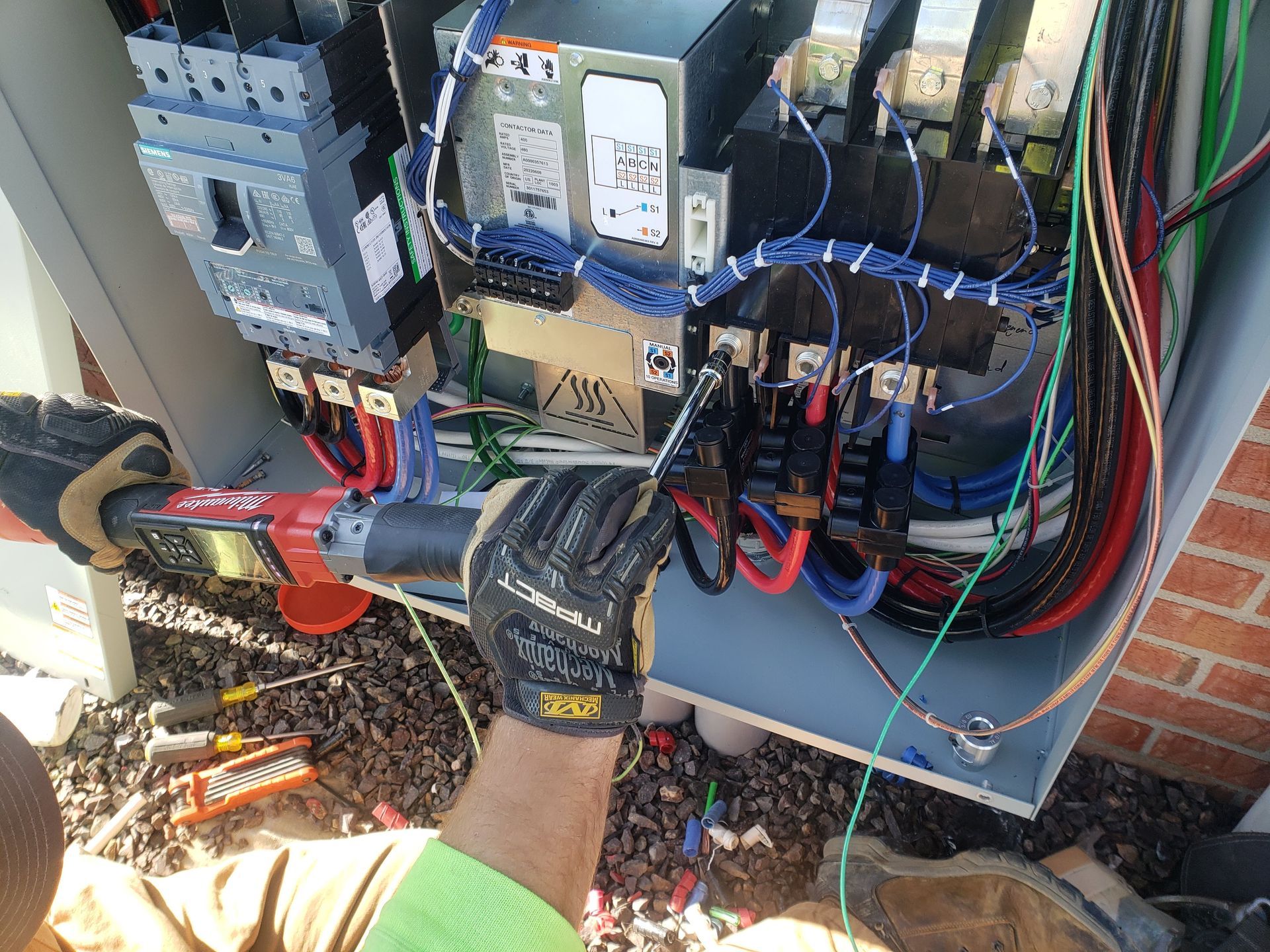 Electrician working inside an electrical box with a red tool, wearing gloves, in outdoor setting.
