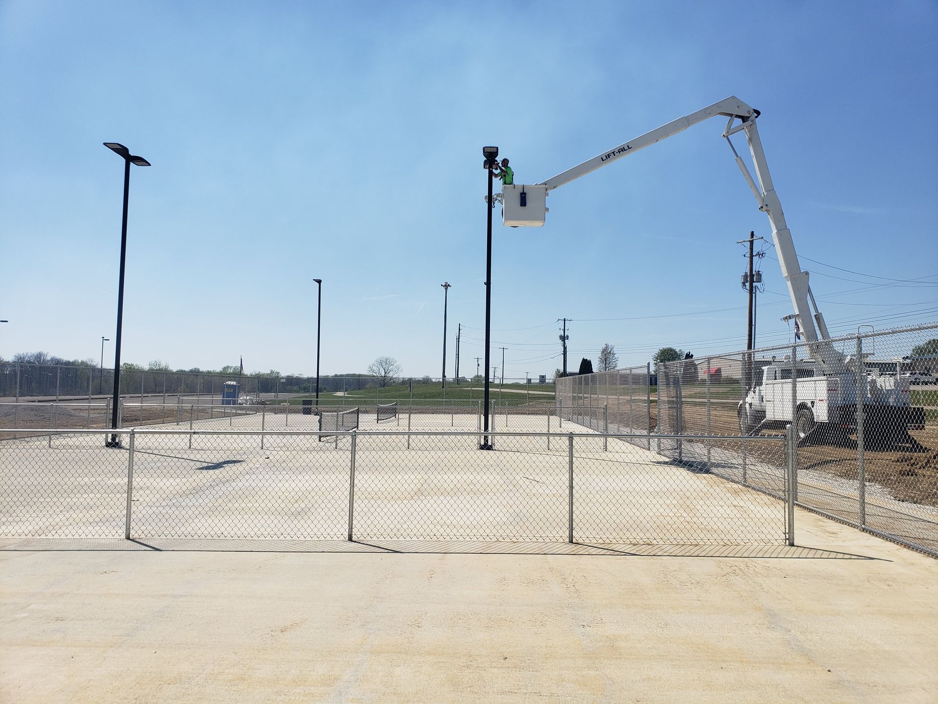 A worker in a lift basket is installing a light fixture on a pole, inside a fenced area with other poles, on a sunny day.