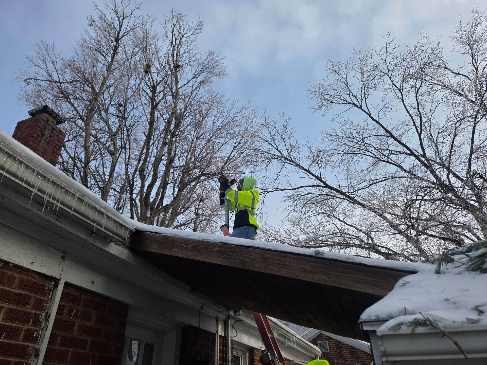 Workers on a snowy roof clearing ice, trees in the background, brick building.