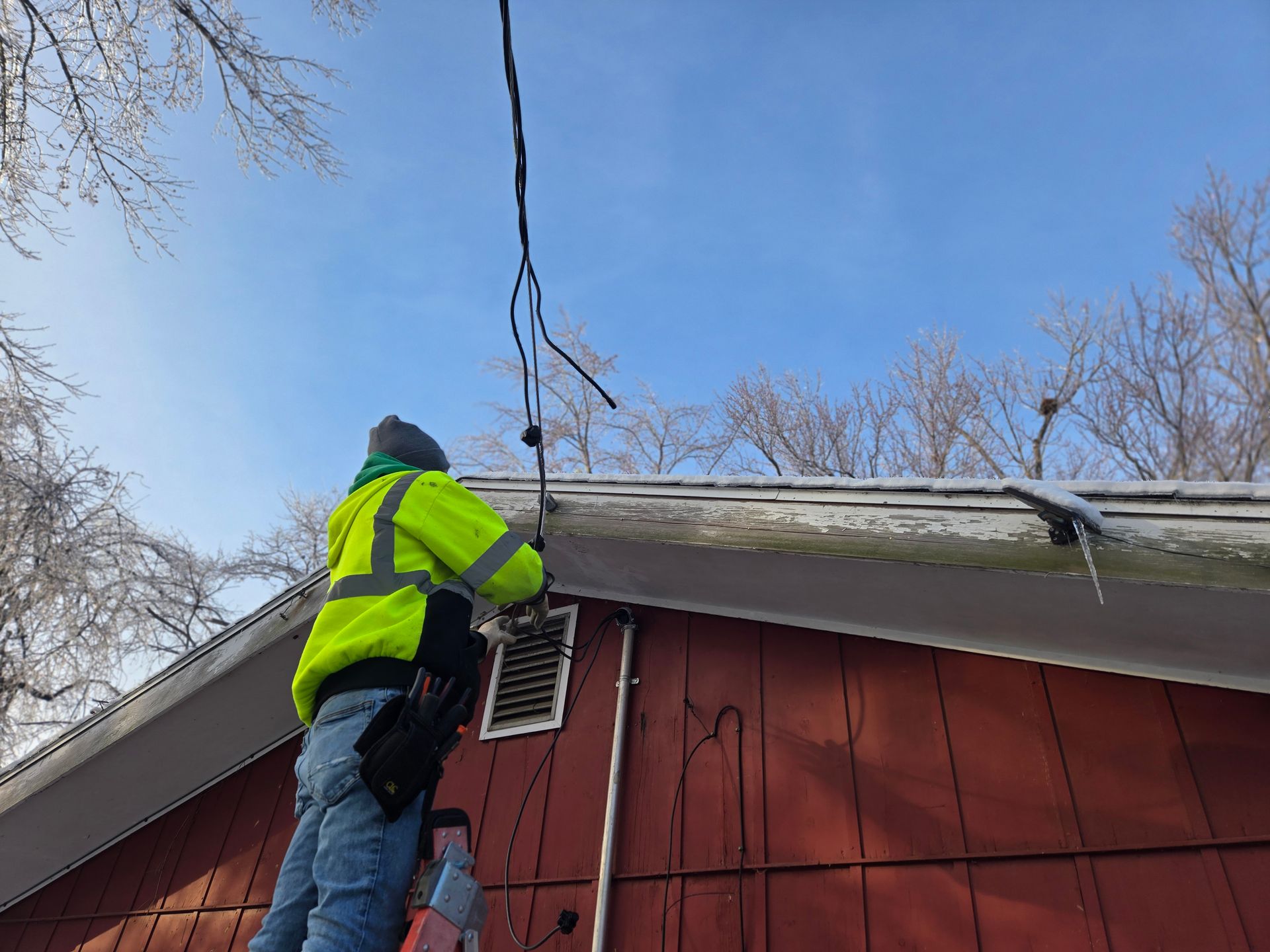 Person in a yellow safety vest on a ladder, working on power lines attached to a red house with a snow-covered roof.