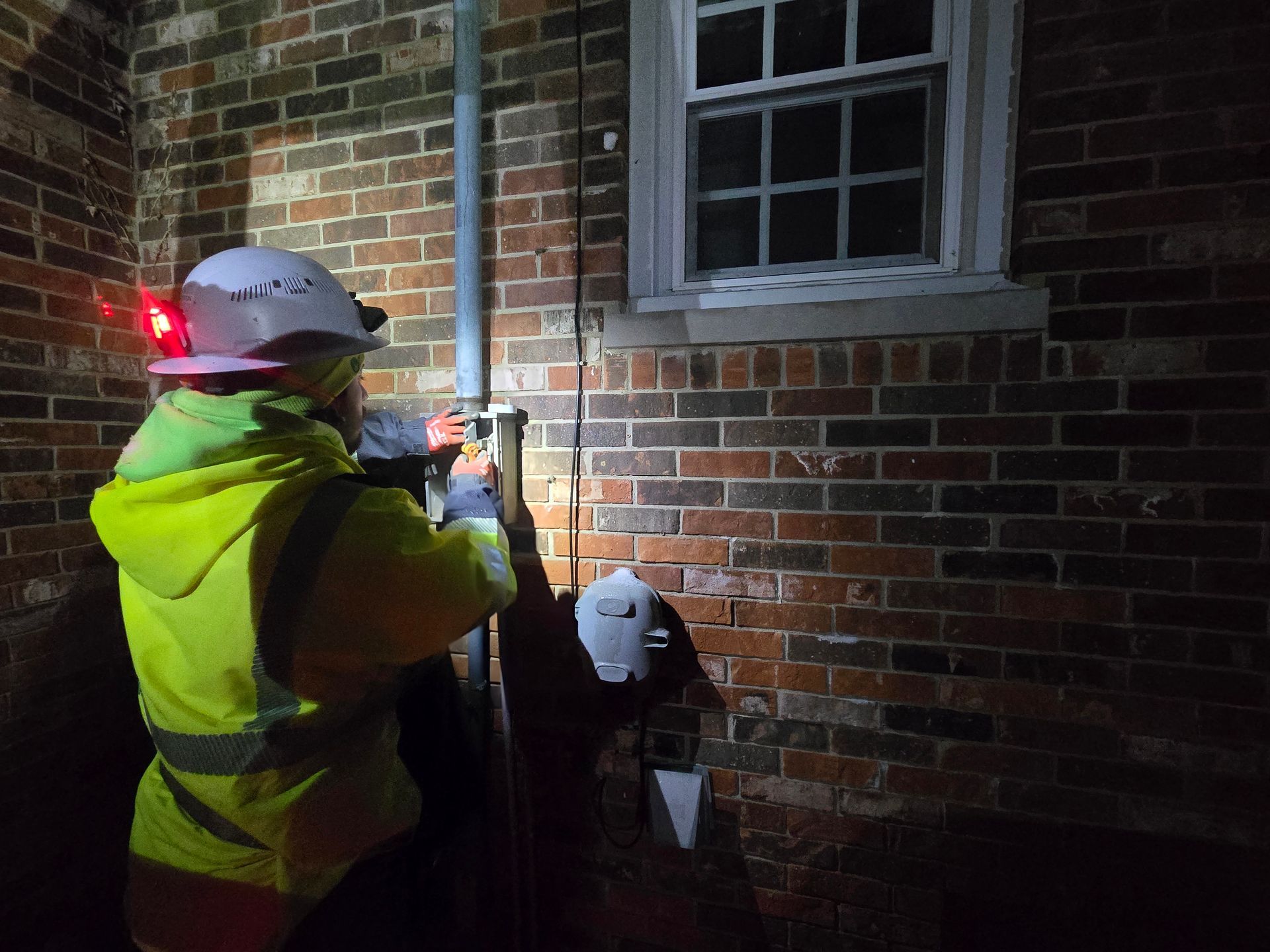 Worker in reflective vest and hard hat working on a pipe attached to a brick wall at night.