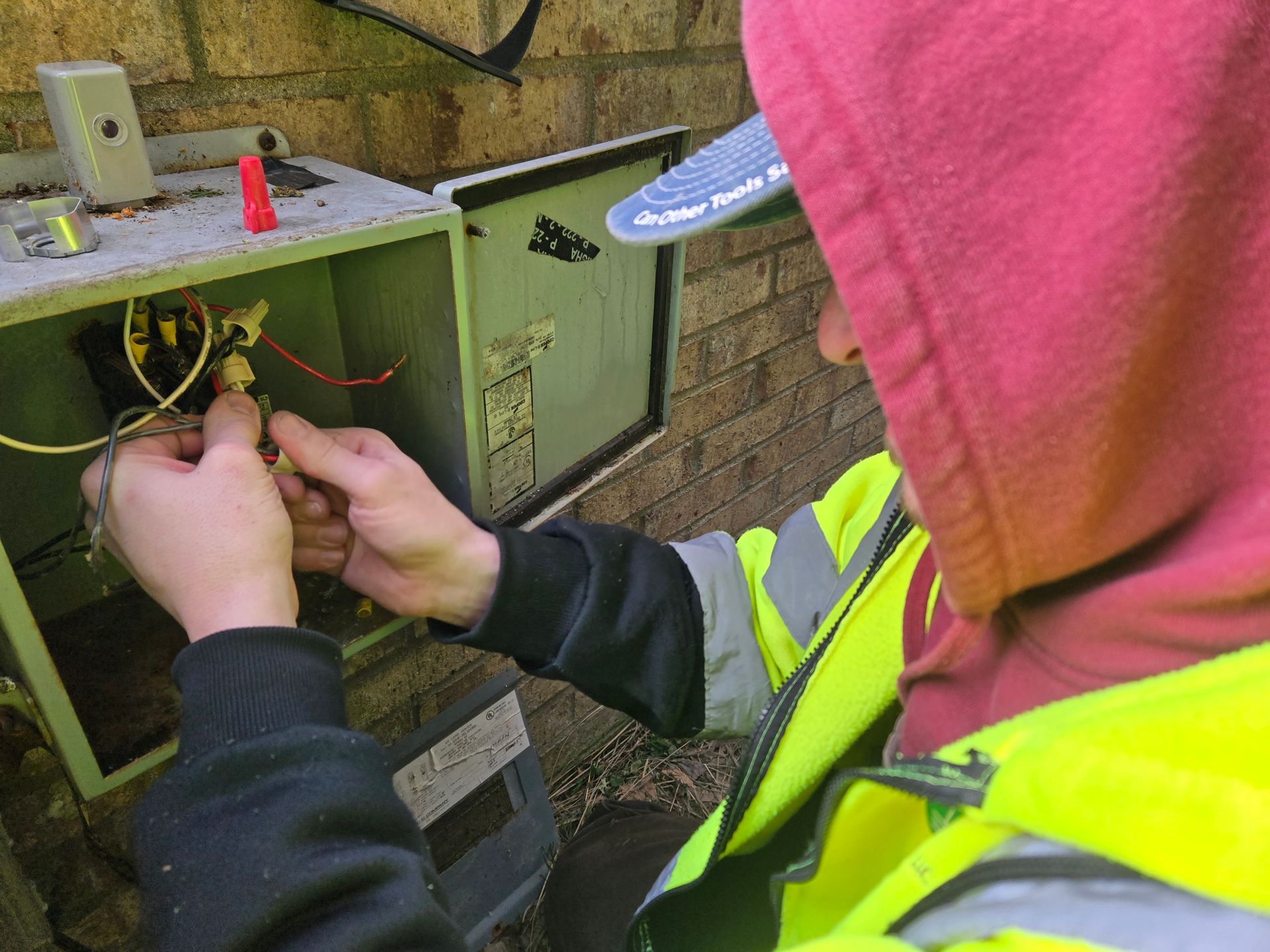 Person in a reflective vest working on electrical wiring in an outdoor box.