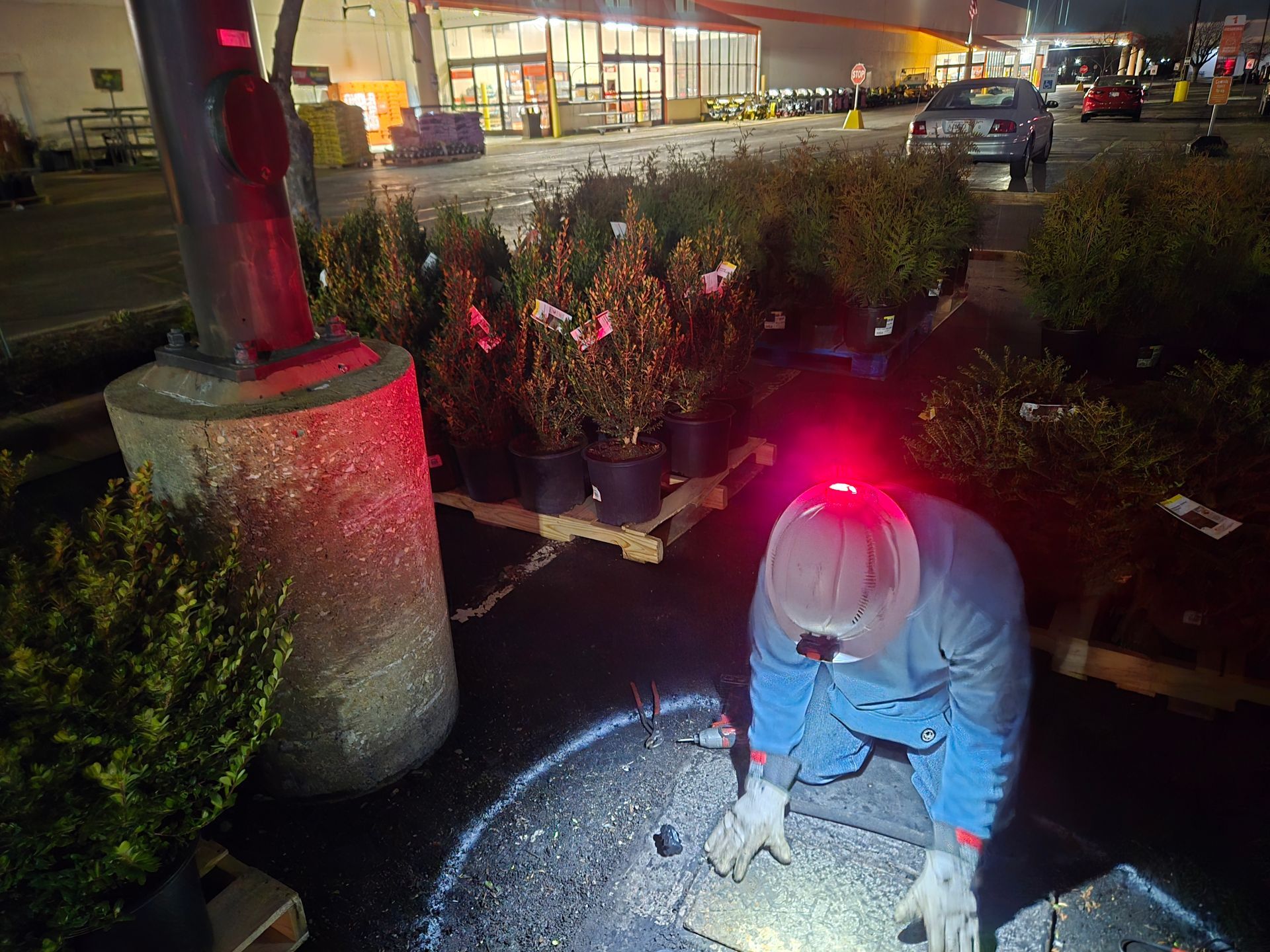 Person working on a ground fixture at a Home Depot, surrounded by plants and illuminated by a headlamp.