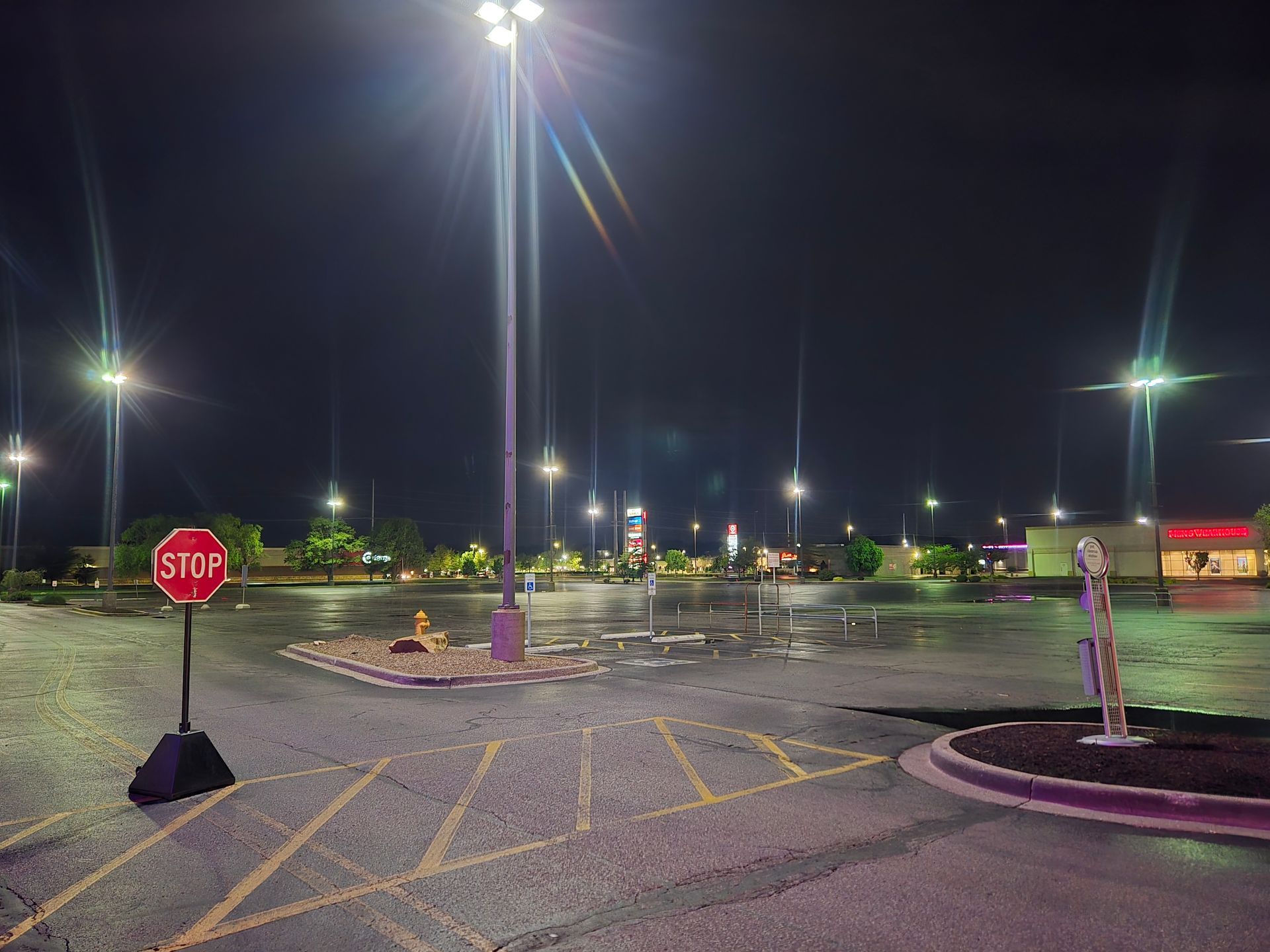 Night view of a wet, empty parking lot under bright streetlights. Stop sign in the foreground.
