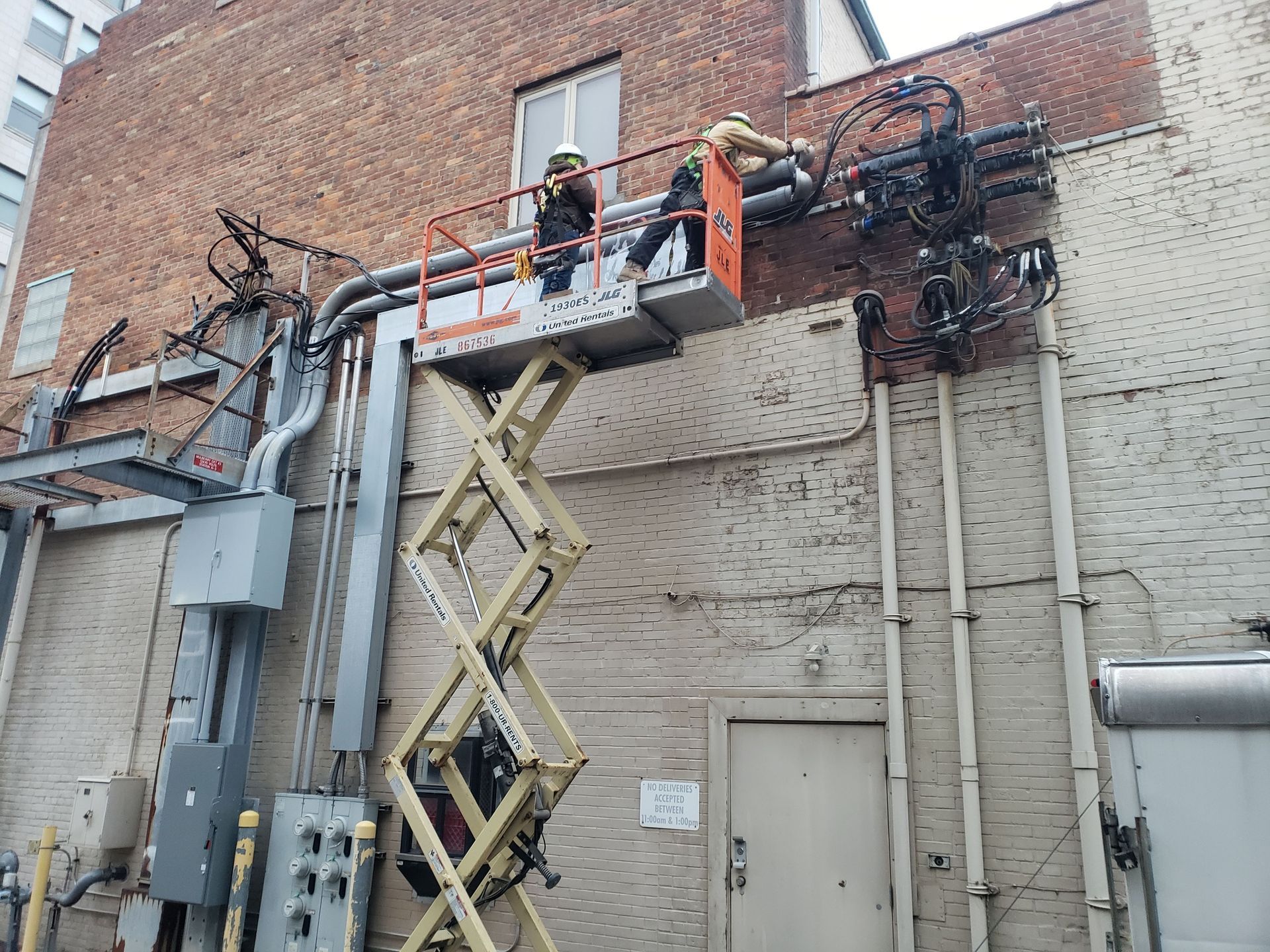 Workers in lift platform repairing electrical equipment on brick building.