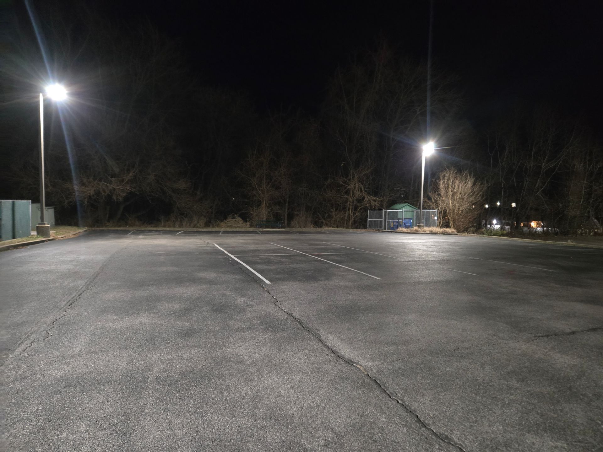 Empty asphalt parking lot at night, illuminated by bright overhead lights. Trees line the background.