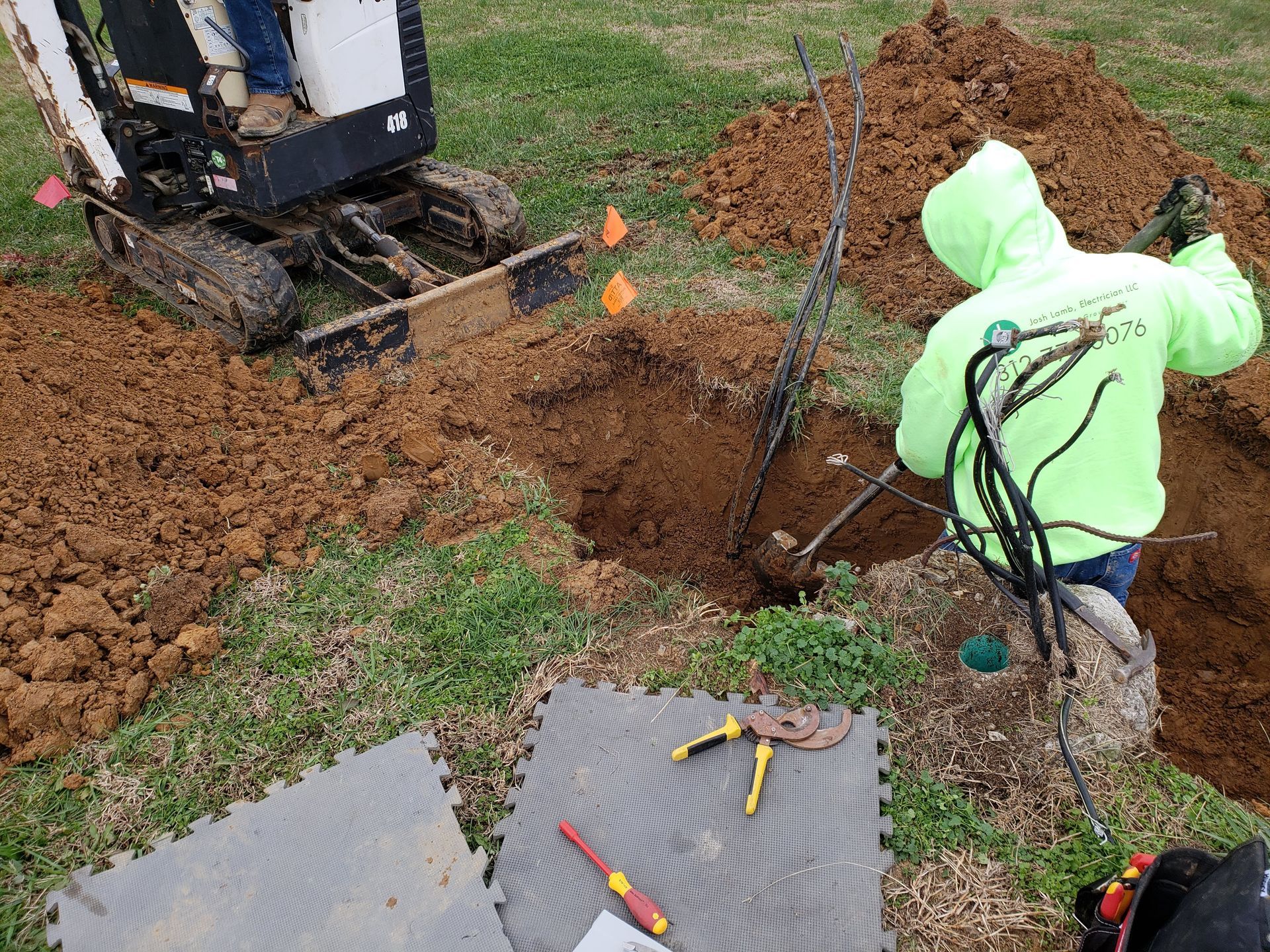 Man in neon green jacket working on wires in a dug hole. Bobcat and tools are nearby on grass.