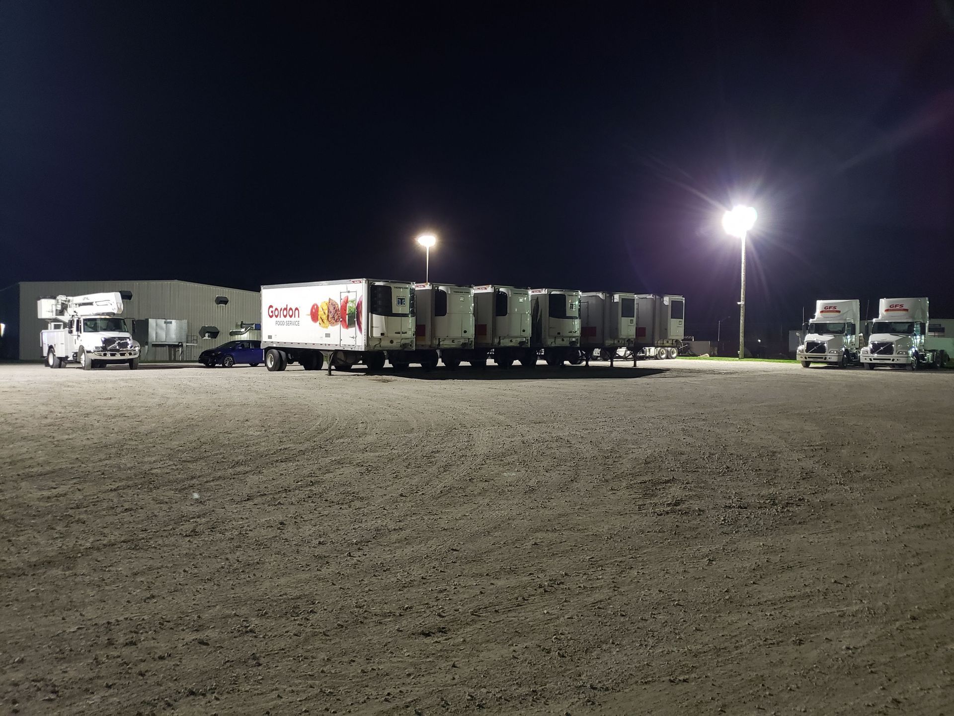 Night scene: trucks and trailers parked on a gravel lot near a building under bright streetlights.