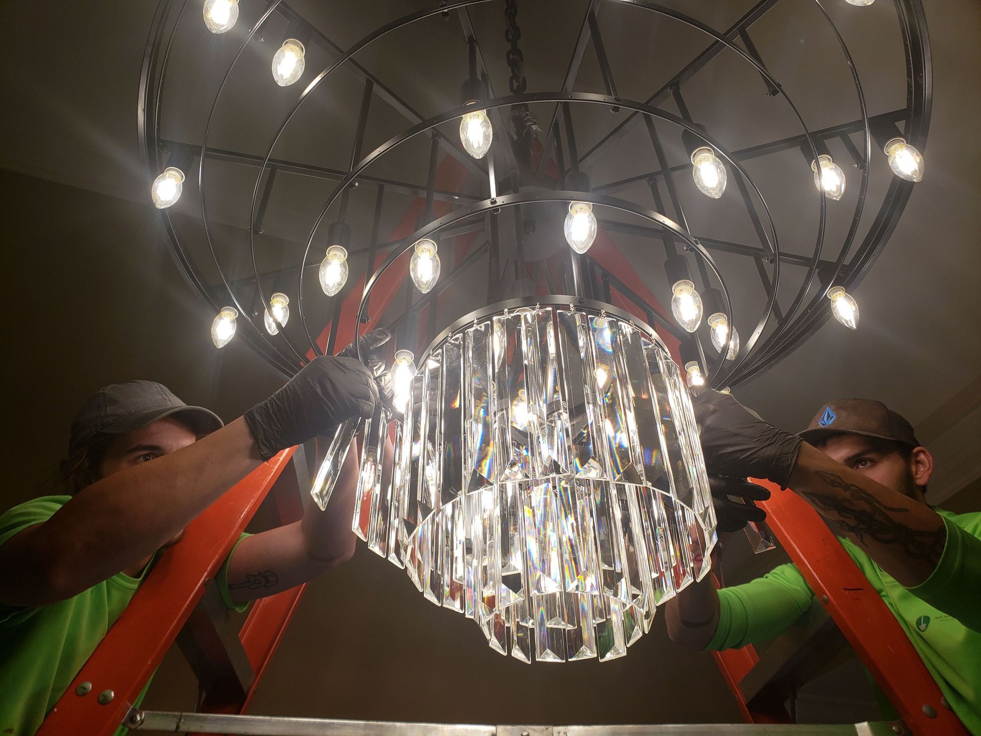 Two workers on ladders installing a large crystal chandelier in a room.