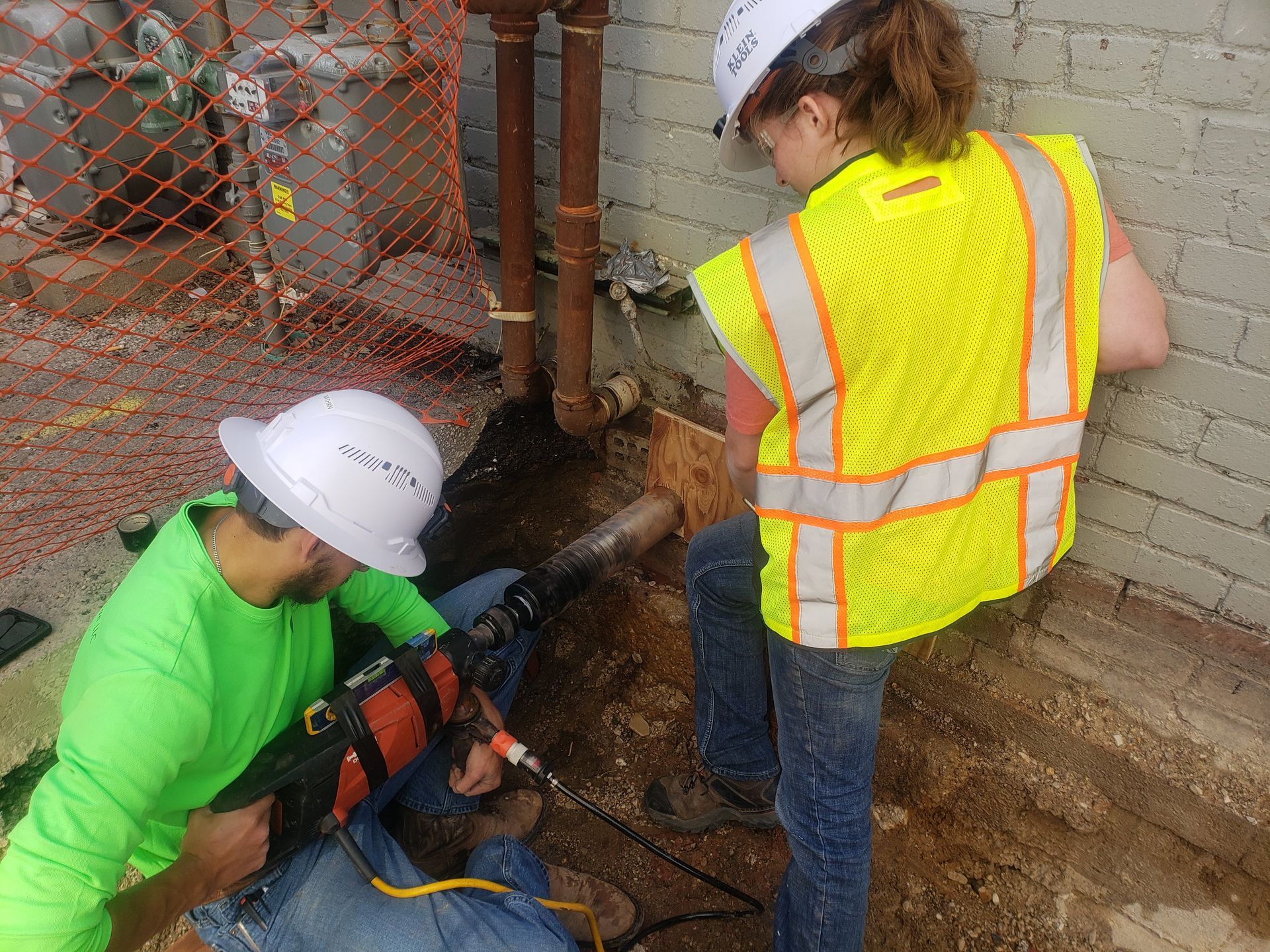 Two construction workers, one drilling, the other observing. Both wearing hard hats and safety vests; working outdoors.