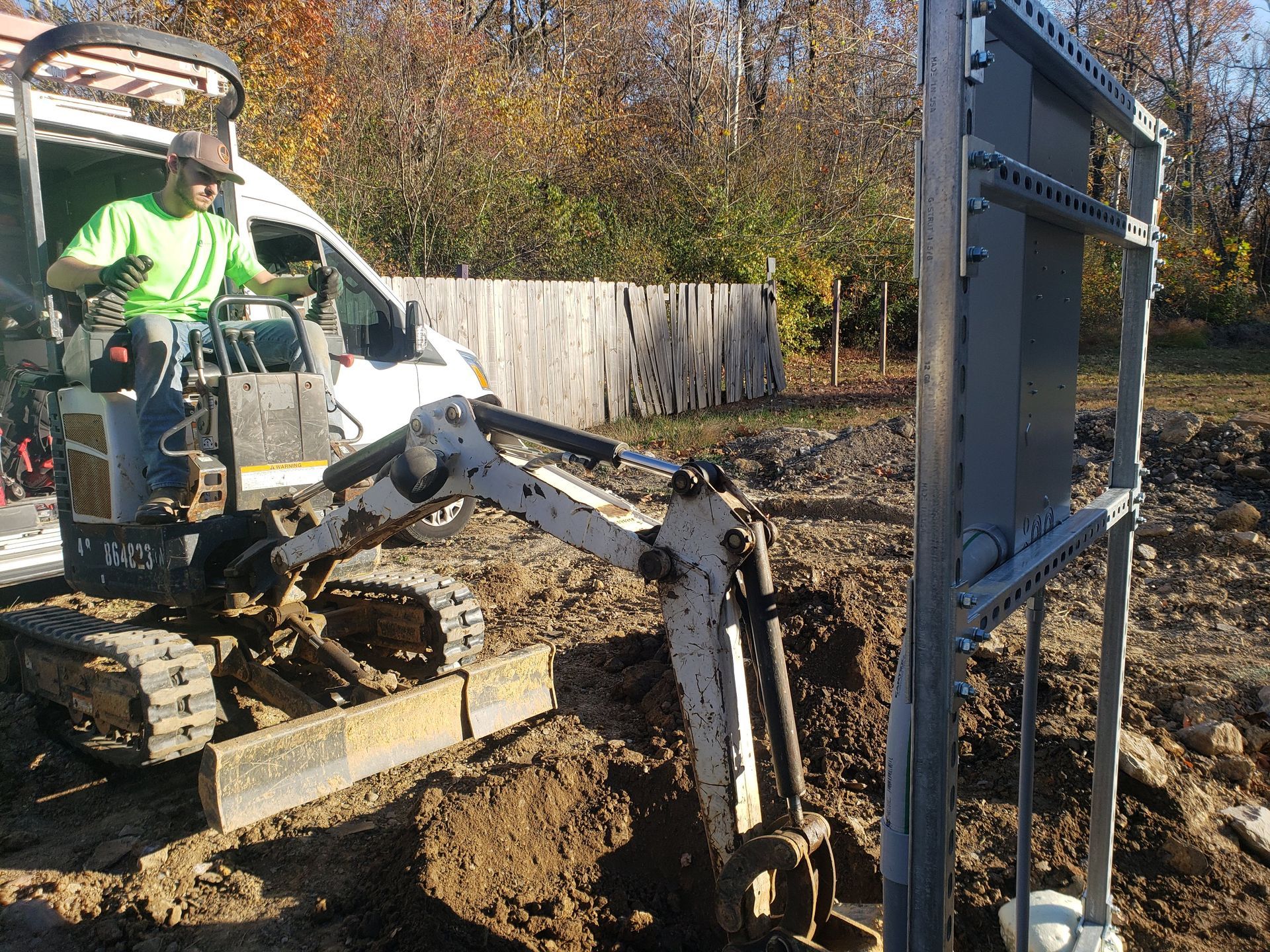 A person operating a mini excavator digging near a metal structure outdoors.