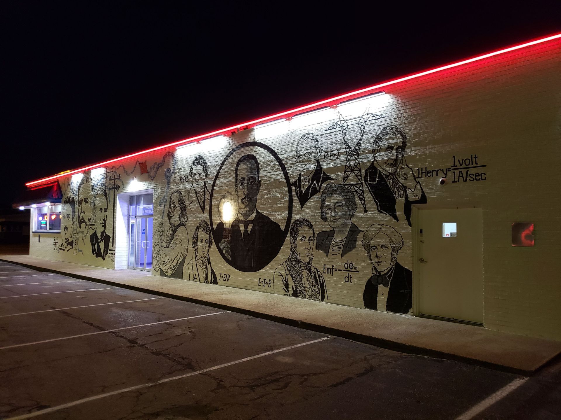 Mural of historical figures on a building at night, lit by red lights. Parking lot in foreground.