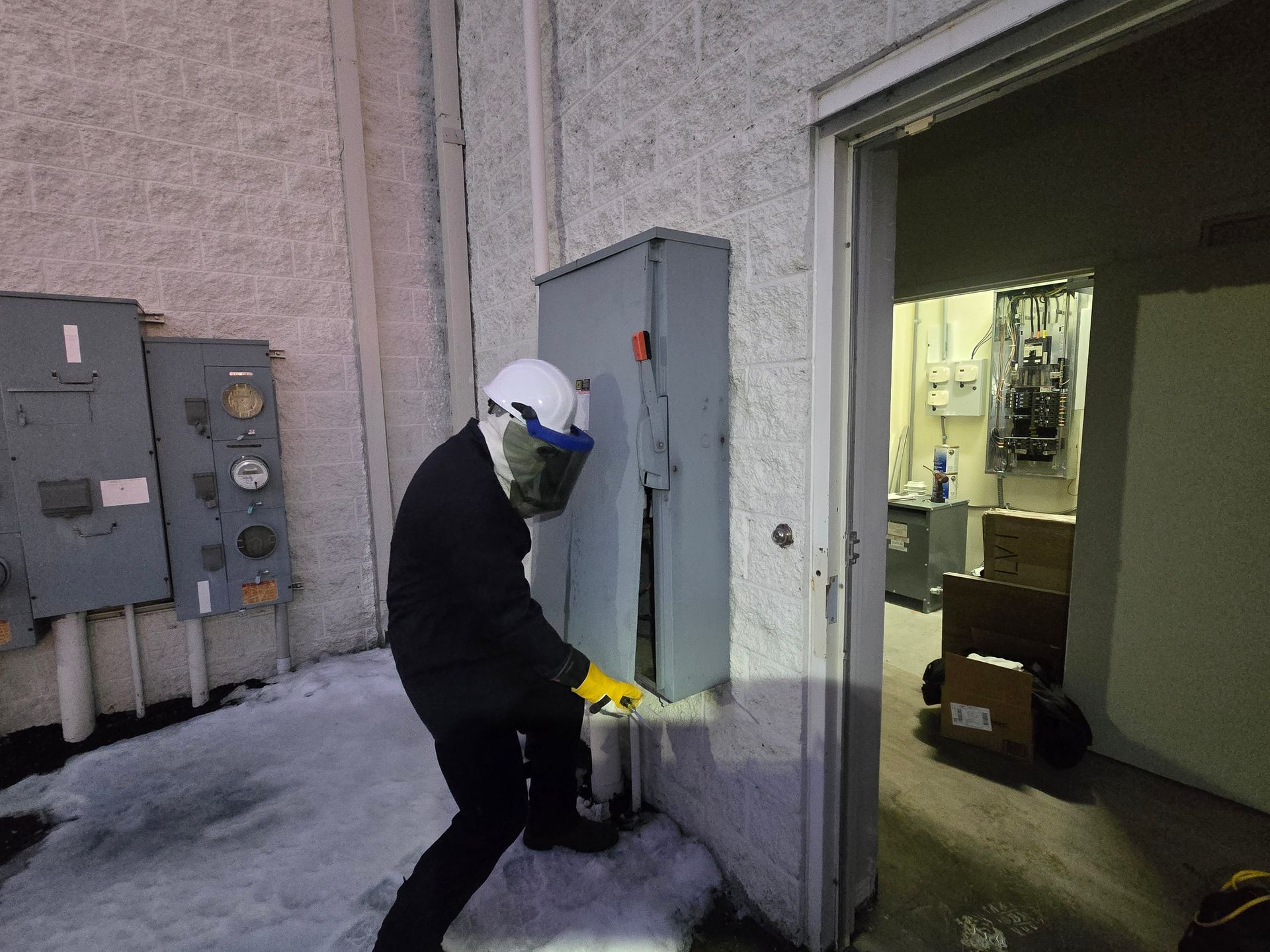 Person in helmet and gloves tampering with a metal electrical box on an exterior wall, near an open doorway.