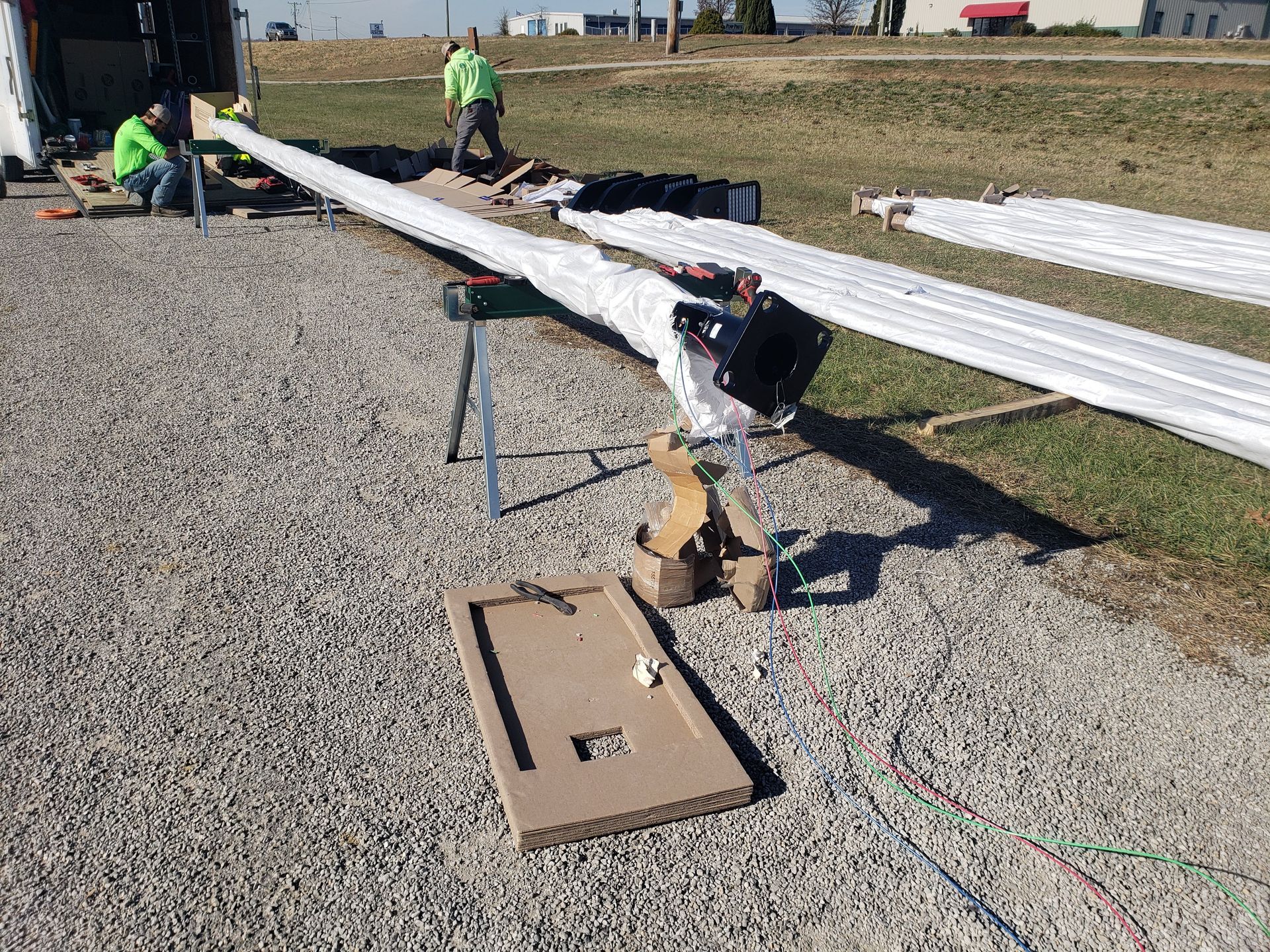 Workers assembling a long, wrapped metal pole outdoors on gravel, with other poles in the background.