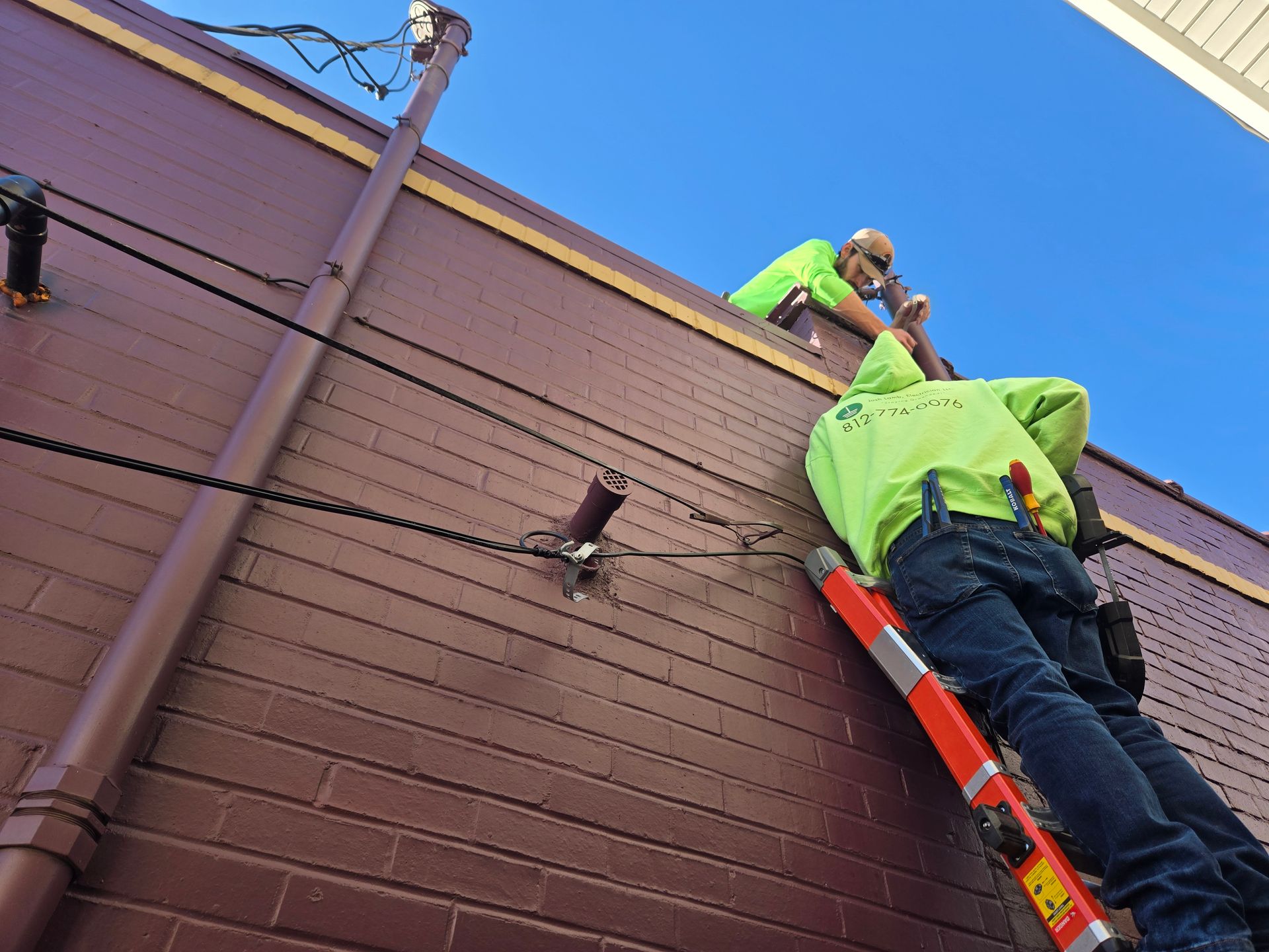 Two workers in safety vests on a ladder and roof fixing power lines on a brick building under a blue sky.