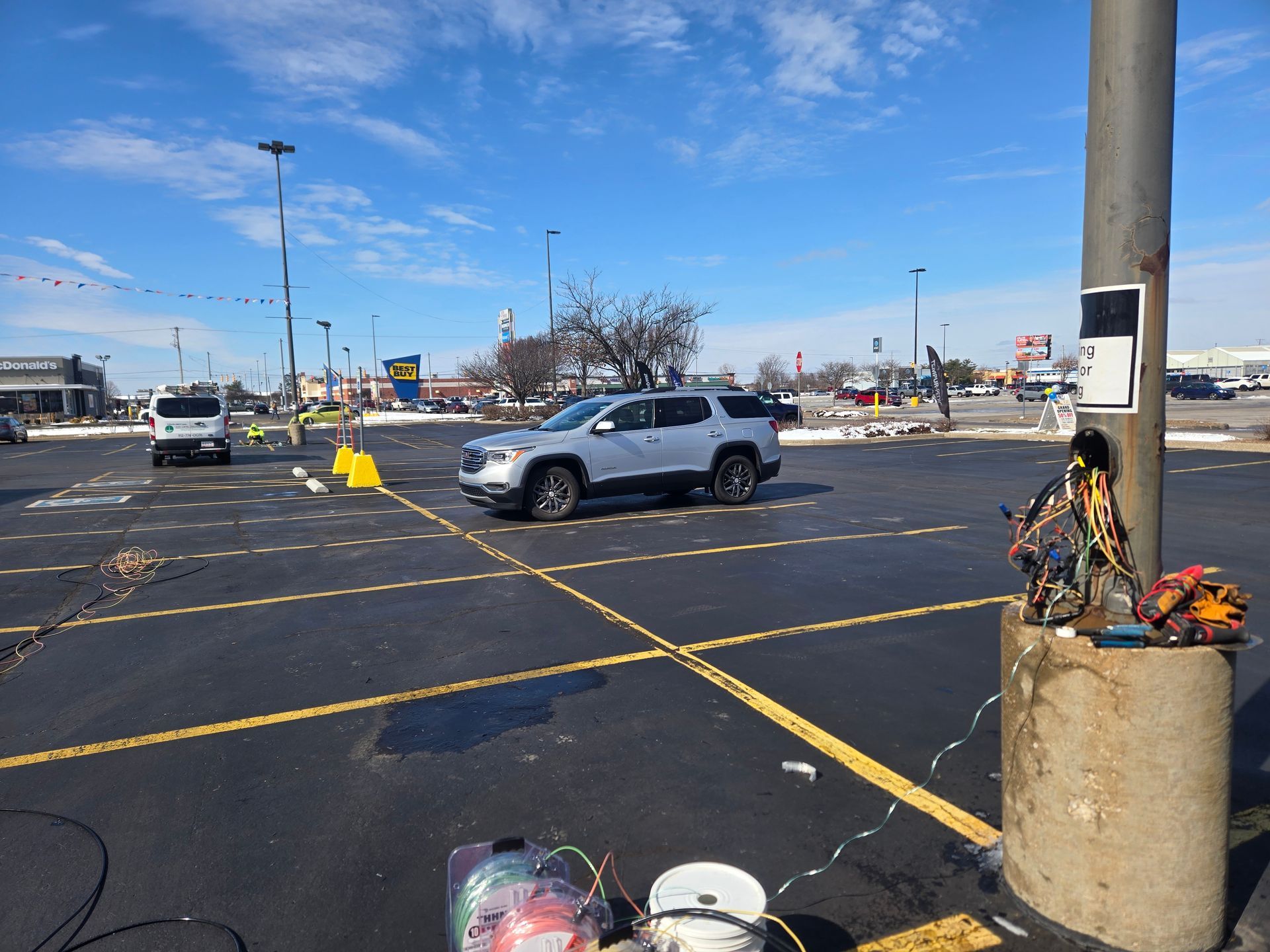 Silver SUV driving in a parking lot on a sunny day. Concrete pole with items next to it.