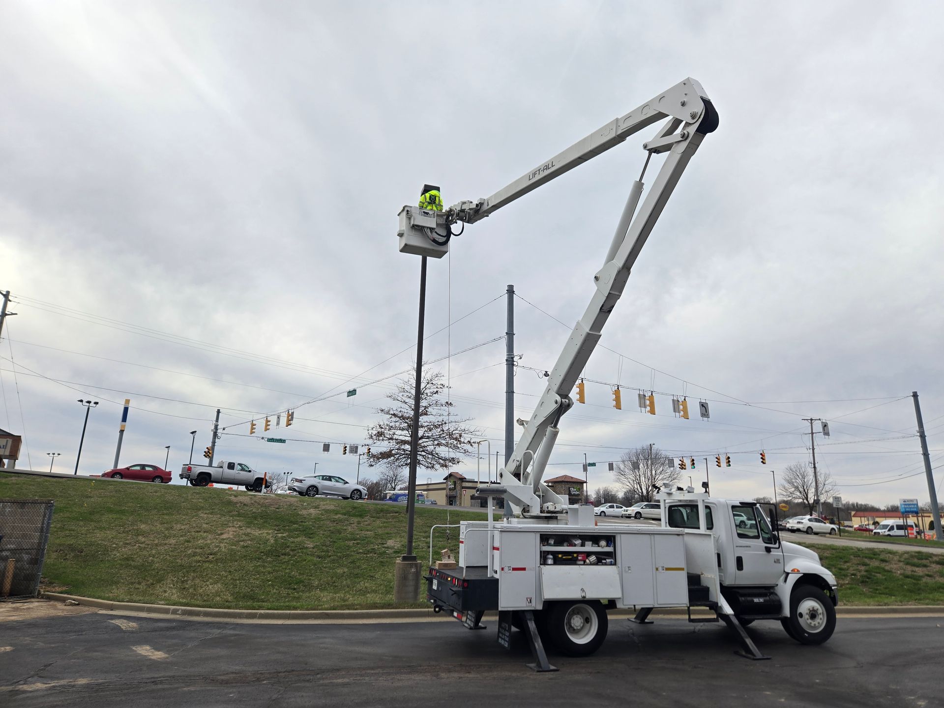 A utility worker in a bucket truck repairs a light pole. Cloudy sky with cars parked in the background.