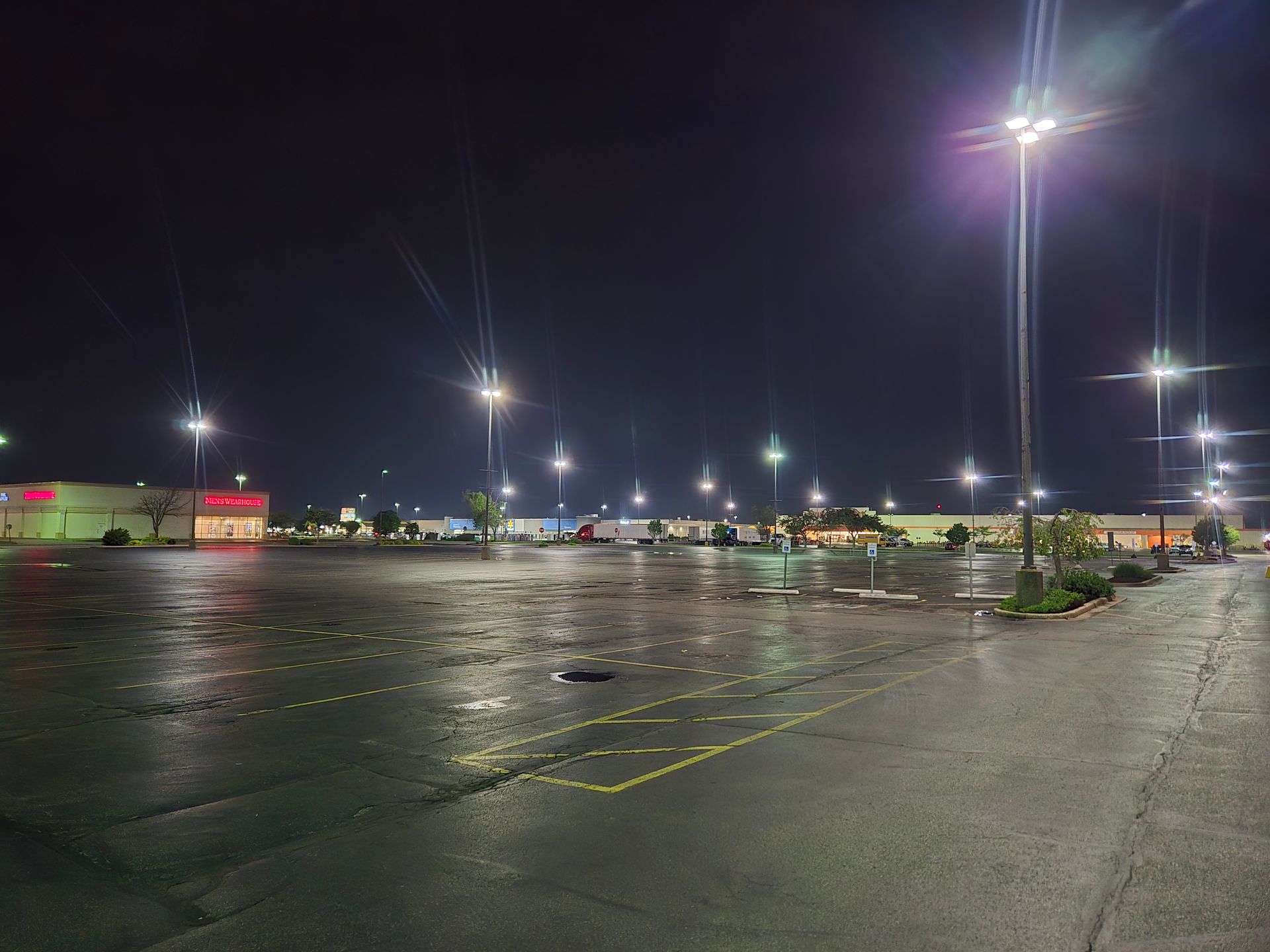 Empty parking lot at night, illuminated by streetlights, with several businesses visible in the background. Wet pavement.