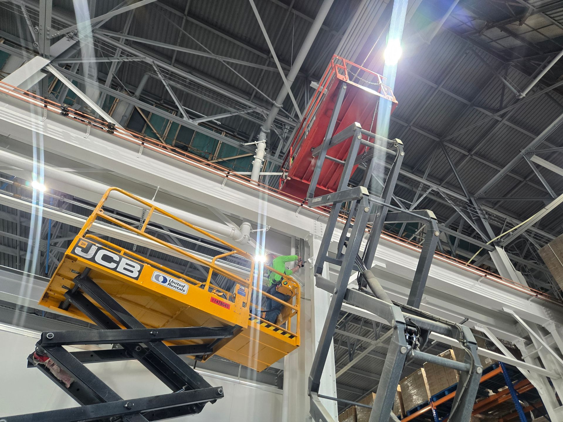 A worker in a yellow JCB lift platform works near a tall metal structure inside a building.