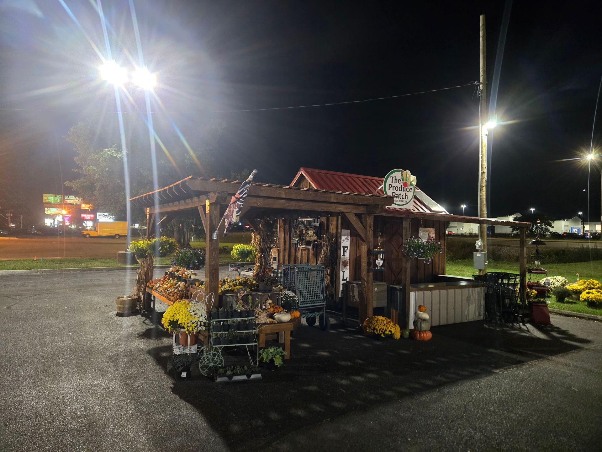 Flower stand at night, displaying mums, pumpkins, and decor. A wooden pergola with a red roof.