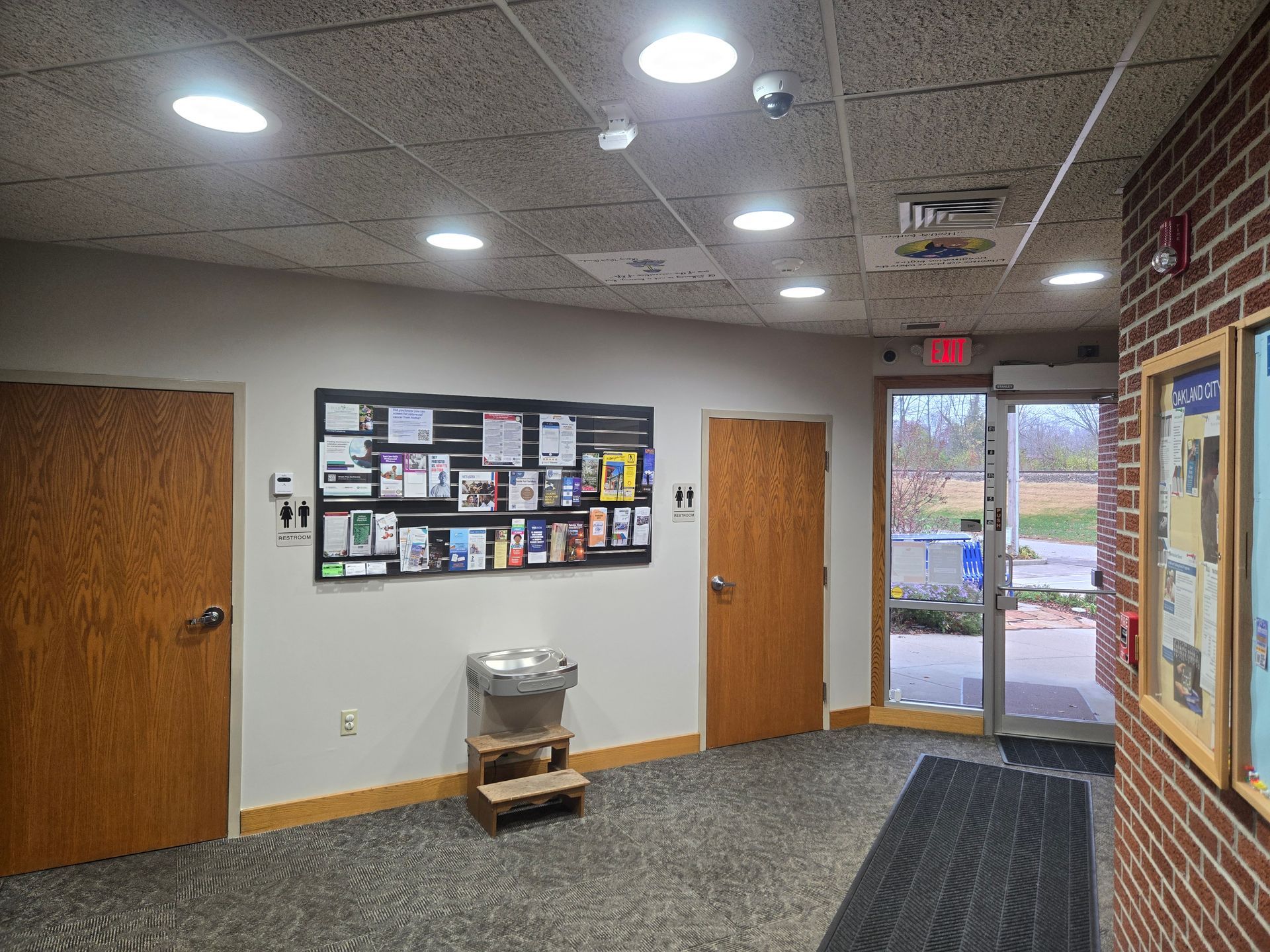Hallway with doors, bulletin board, trash can, and an open doorway to the outside.