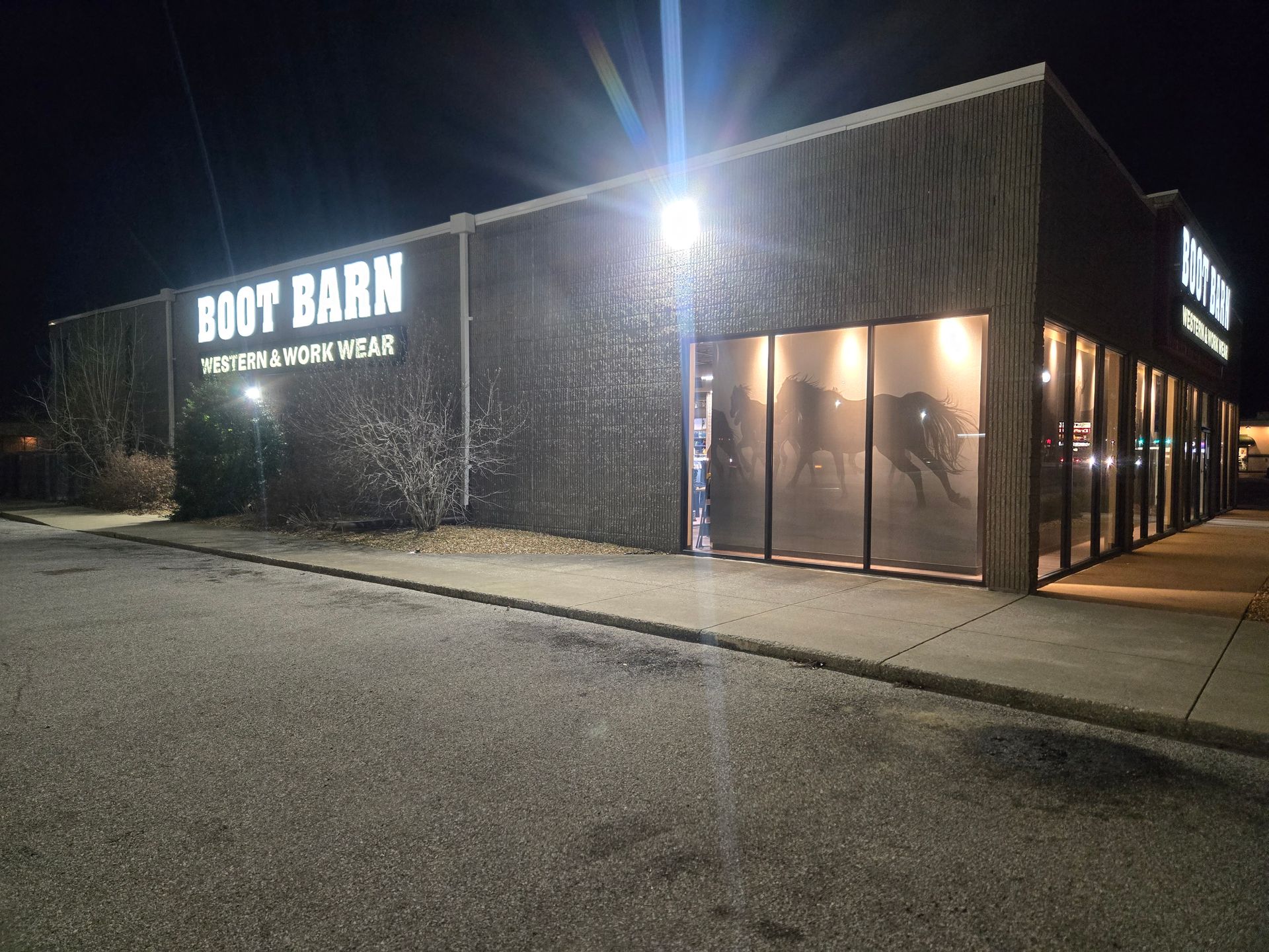 Boot Barn store exterior at night, lit signage, large windows, and parking lot.