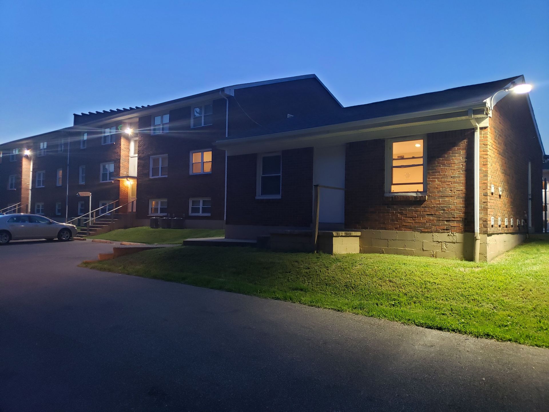 Brick apartment building at dusk, lit by outdoor lights. A car sits in the parking area.