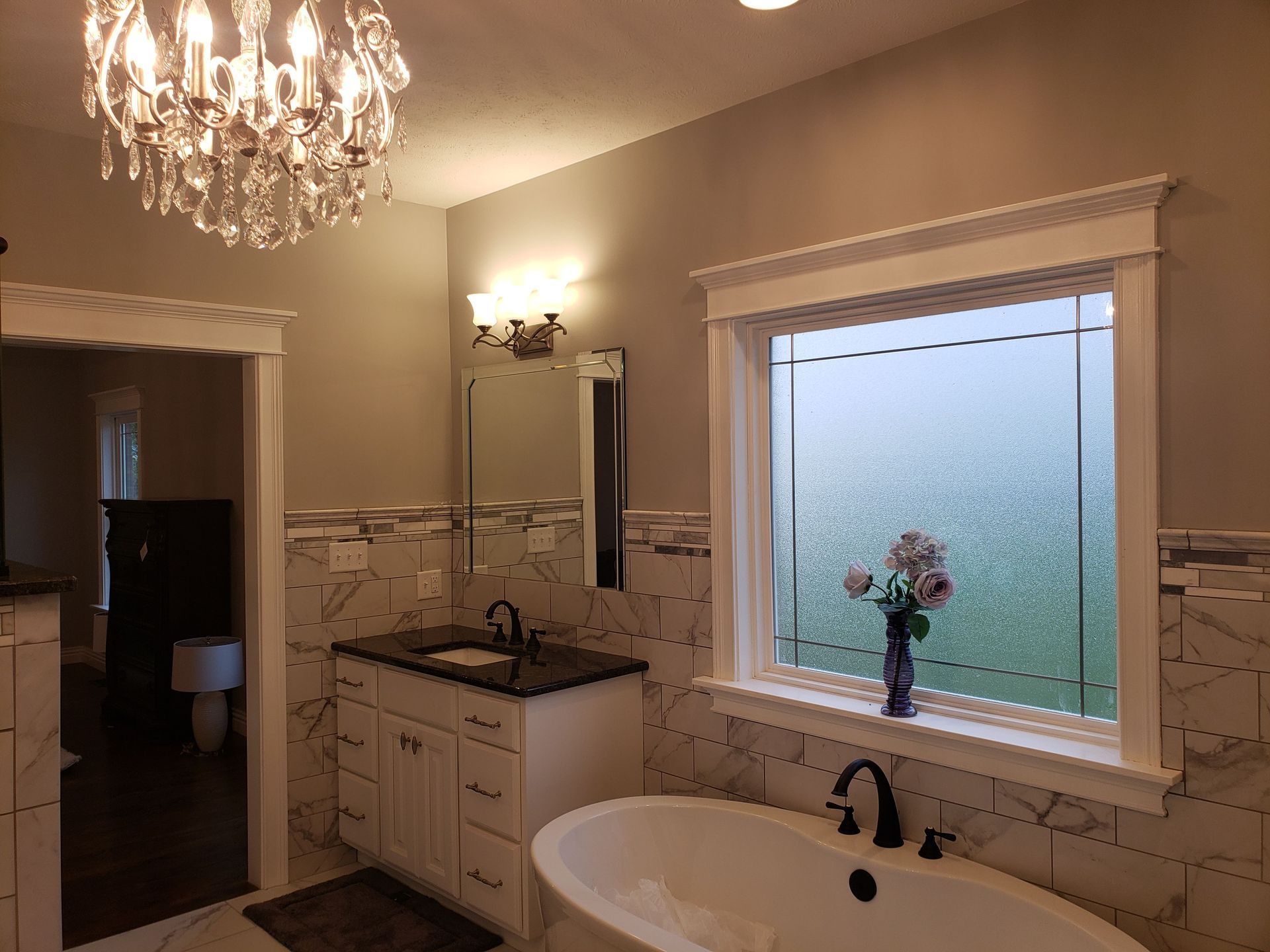 Bathroom with a window, tub, vanity, and crystal chandelier. White and gray tones, modern design.